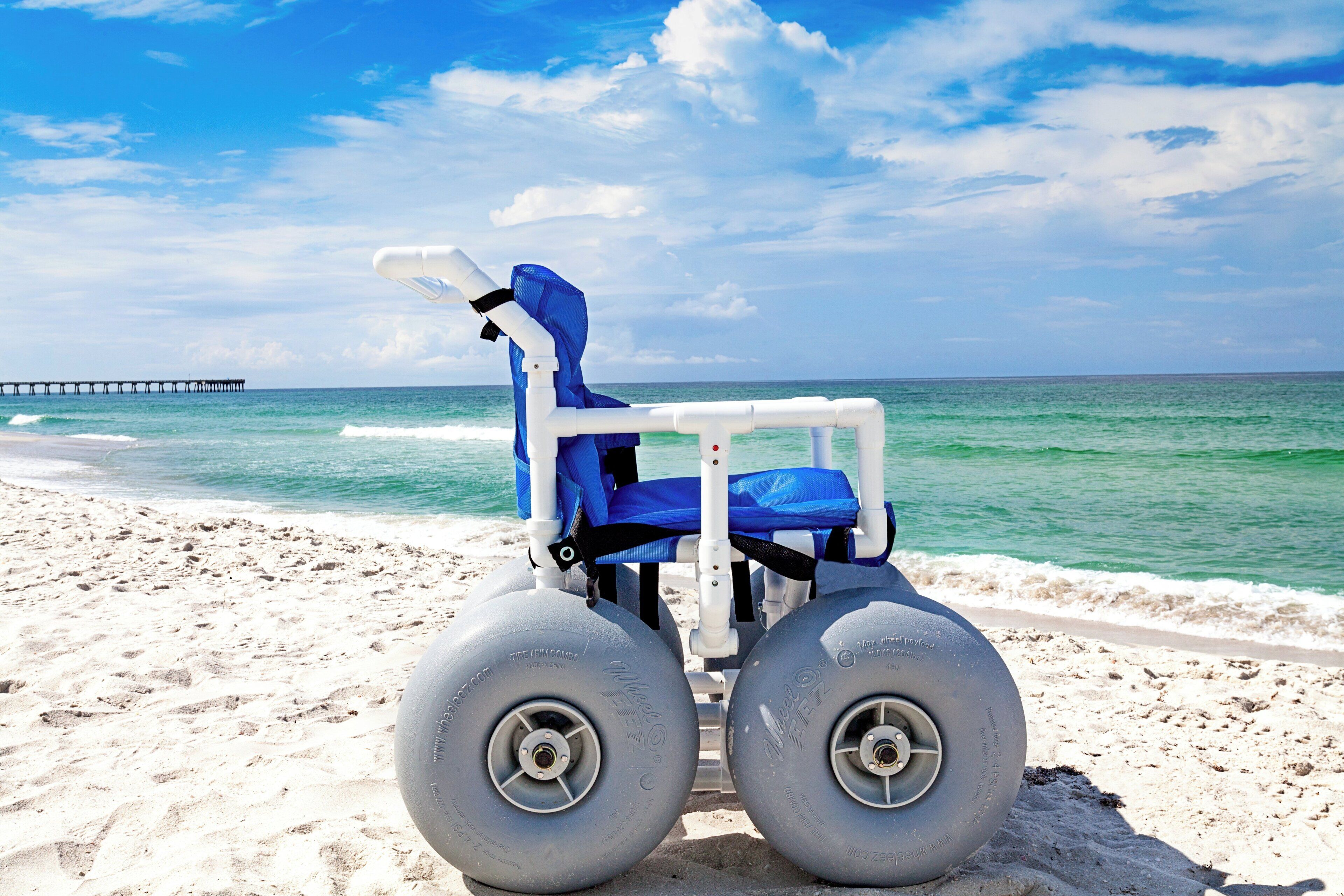 A beach chair with wheels on a sandy beach with a clear sky and a pier in the background.