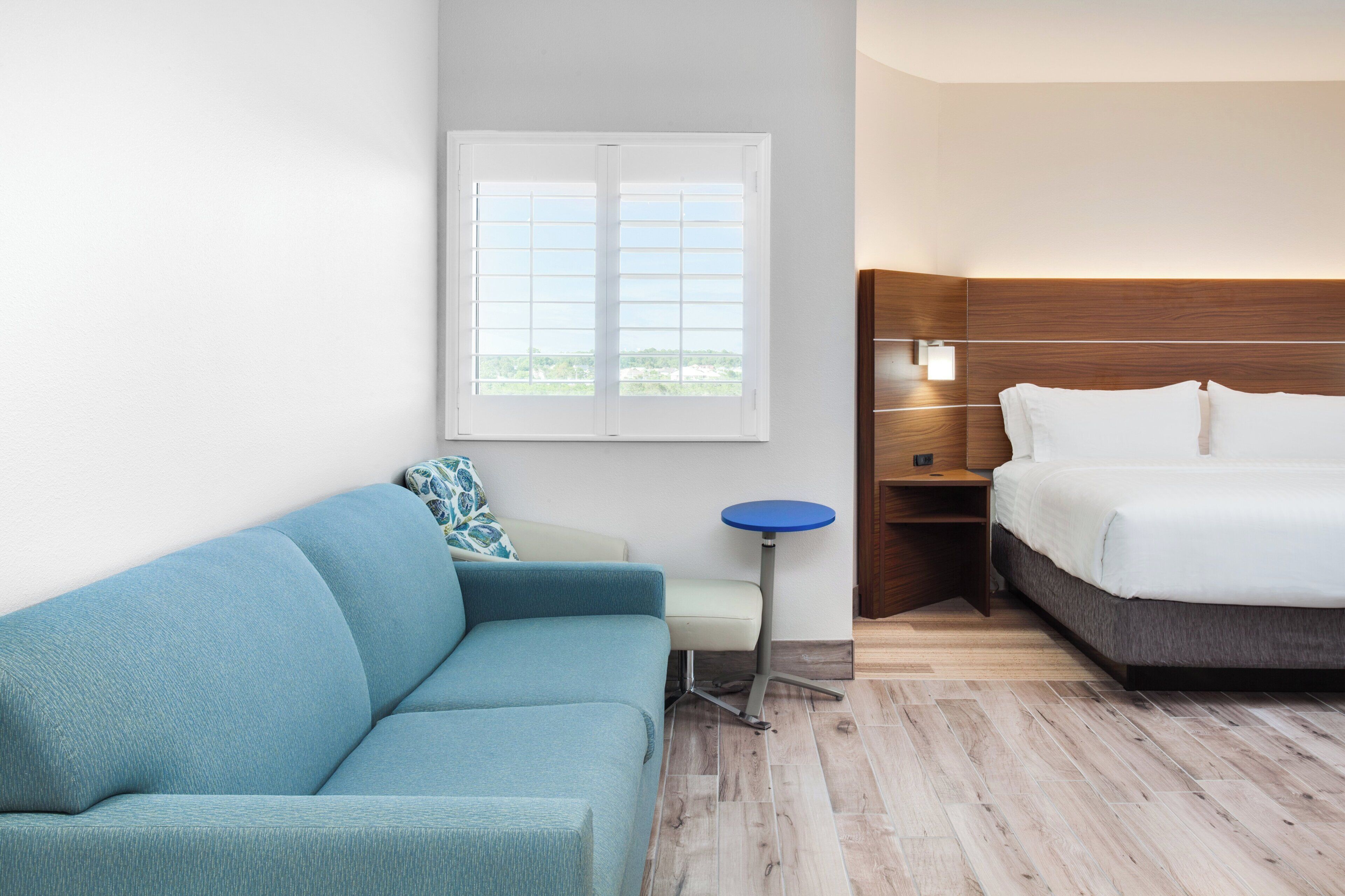 Een hotelkamer met een blauwe bank, een klein rond tafeltje, een bed met wit beddengoed en een raam met uitzicht op groen.