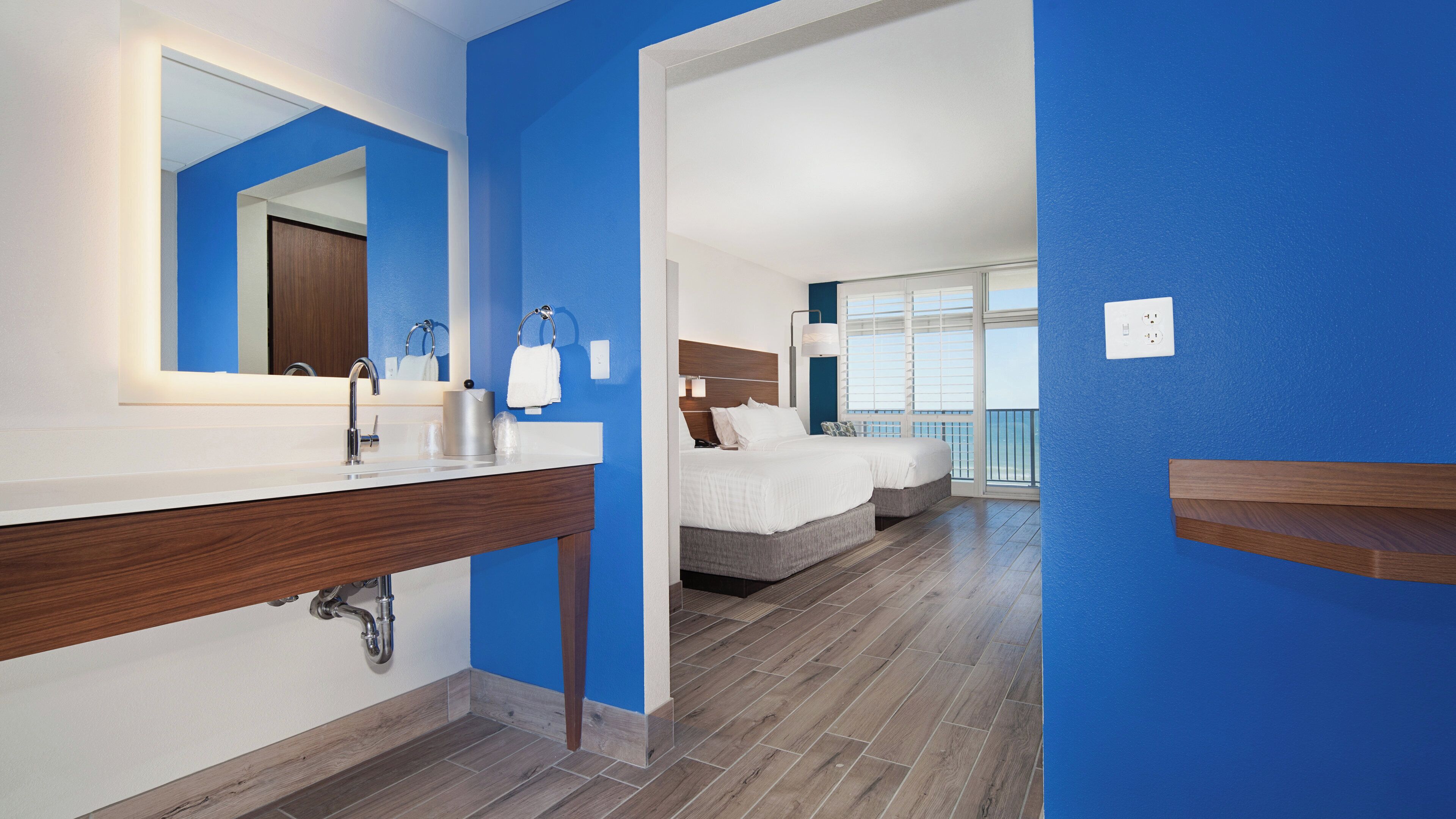 A hotel room with a modern bathroom, a large mirror, a sink, and a bed with a headboard.