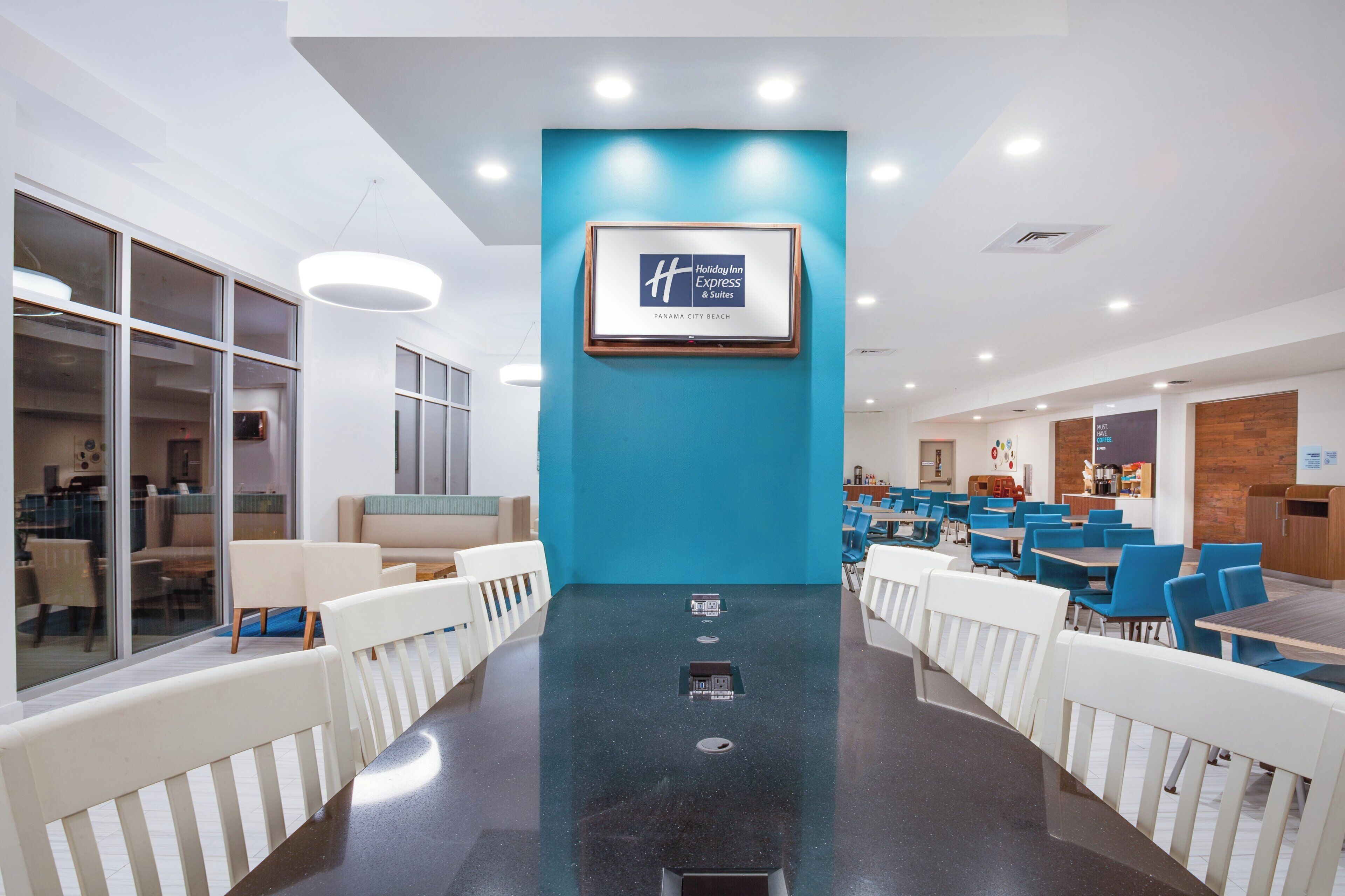 A modern hotel lobby with a central wall-mounted sign, seating areas, and a dining section.