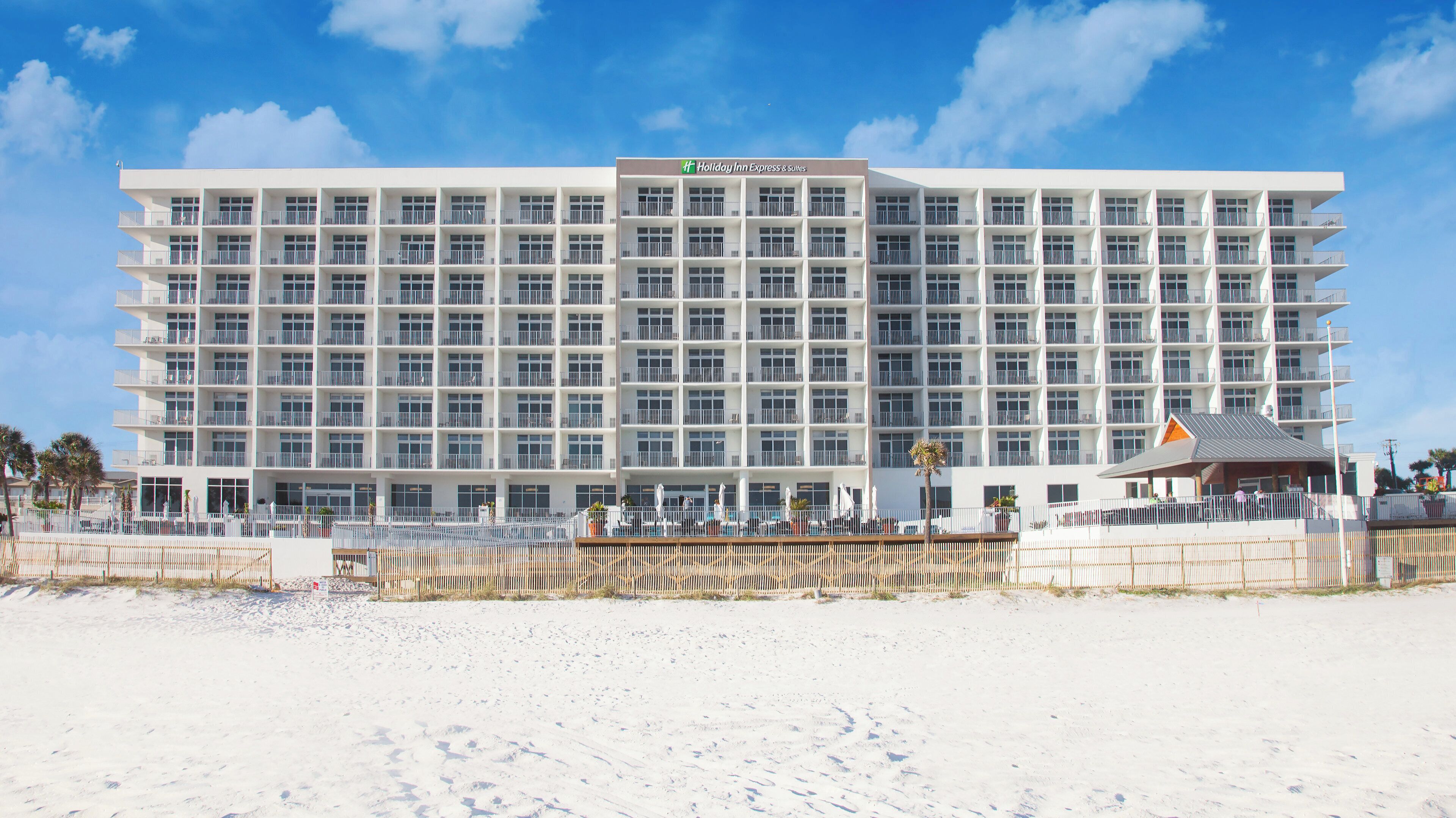 A large, multi-story hotel with a flat roof and numerous balconies, located on a sandy beach.
