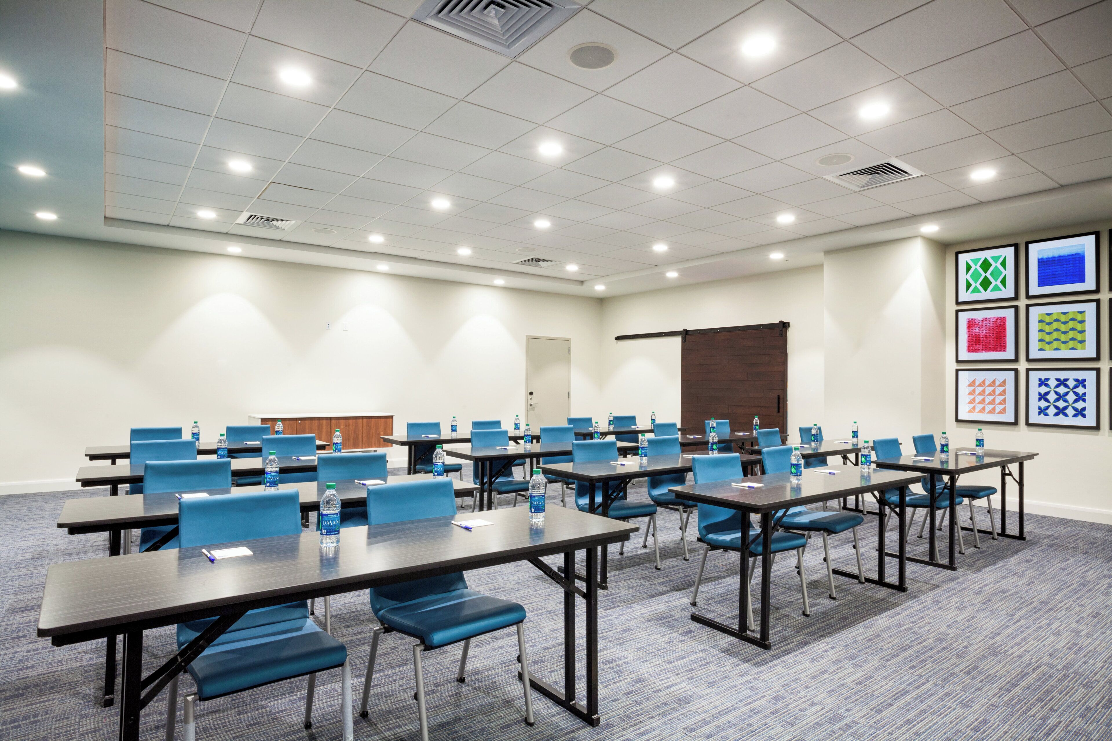 A conference room with multiple tables and chairs, equipped with water bottles, and a projector screen on the wall.