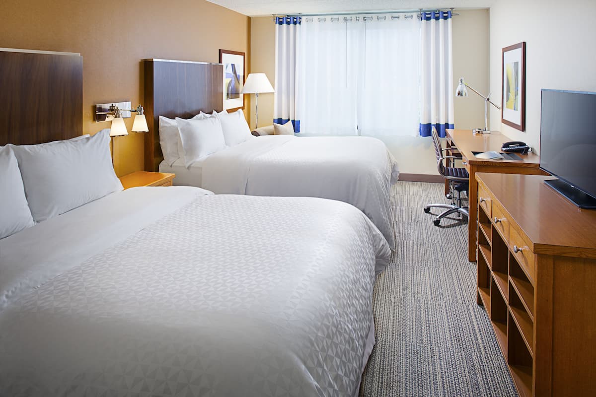 A hotel room with two beds, a desk, a chair, a TV, and a lamp.