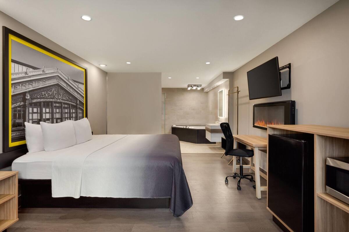 A modern hotel room with a large bed, a desk, a chair, a TV, and a fireplace.
