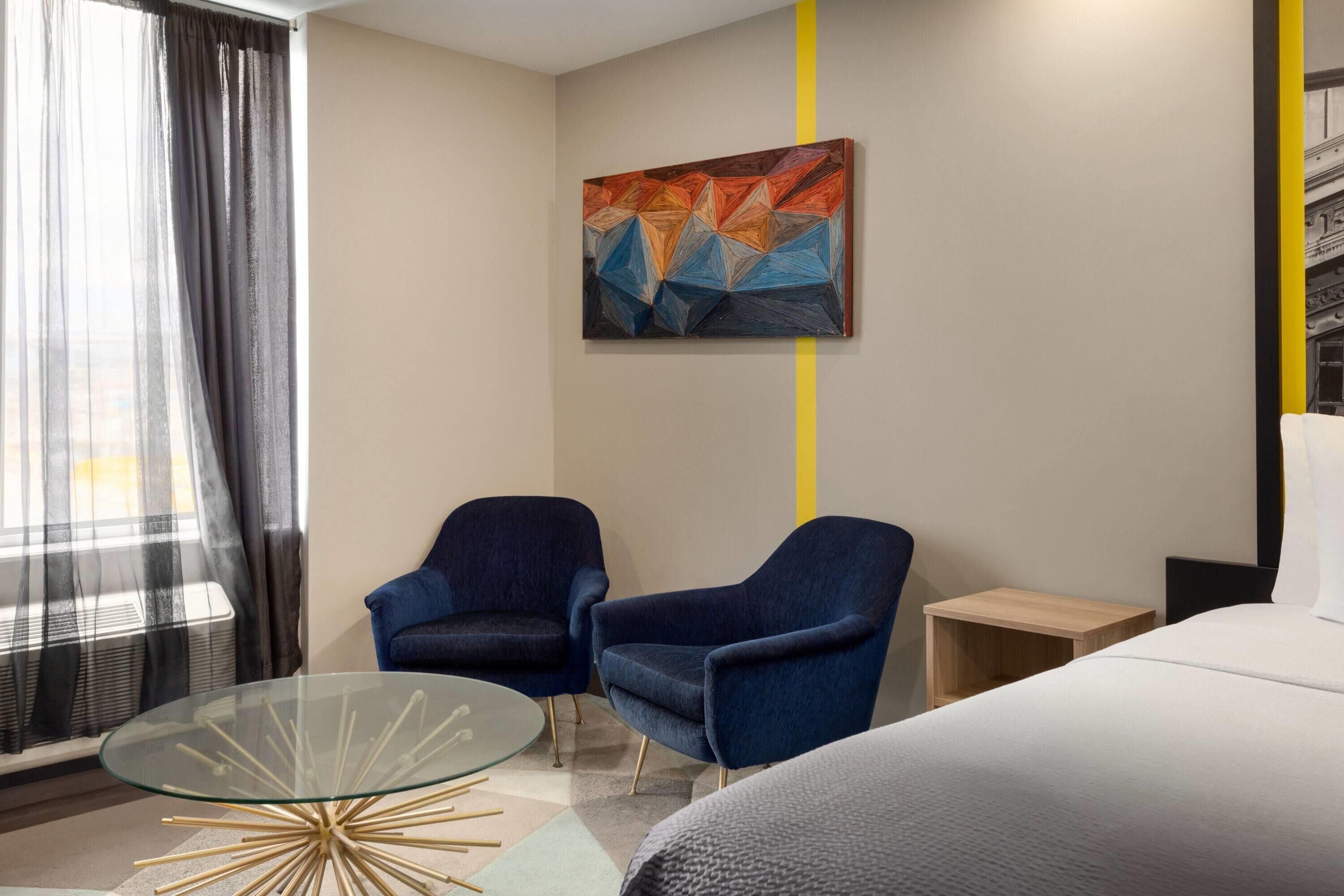 A modern hotel room with a glass coffee table, two blue armchairs, a bed, a small wooden nightstand, and a painting on the wall.