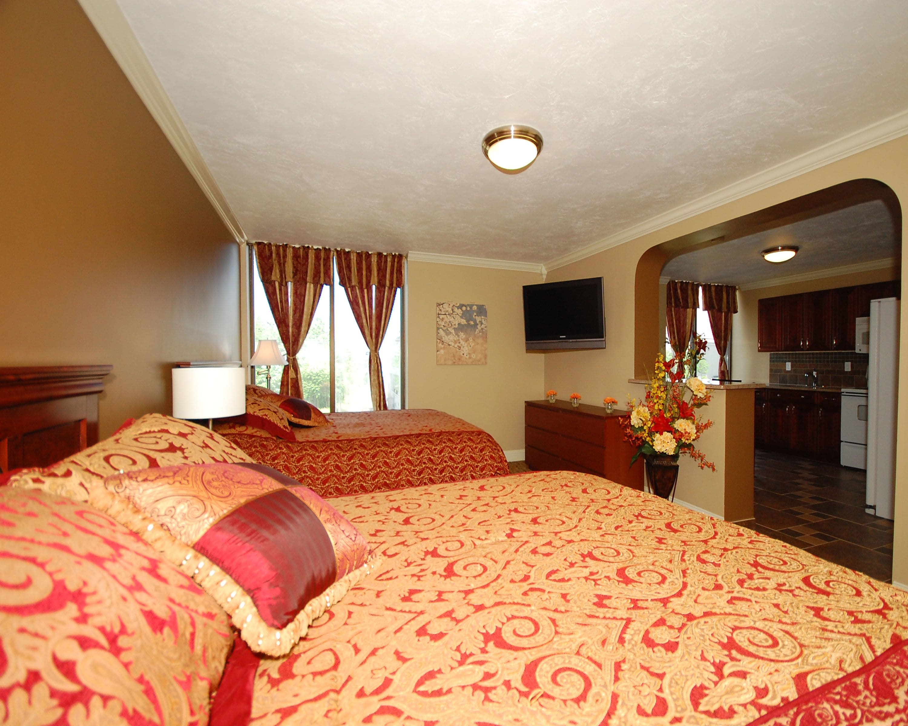 A hotel room with a large bed, a dresser, a TV, and a view of the kitchen and balcony.