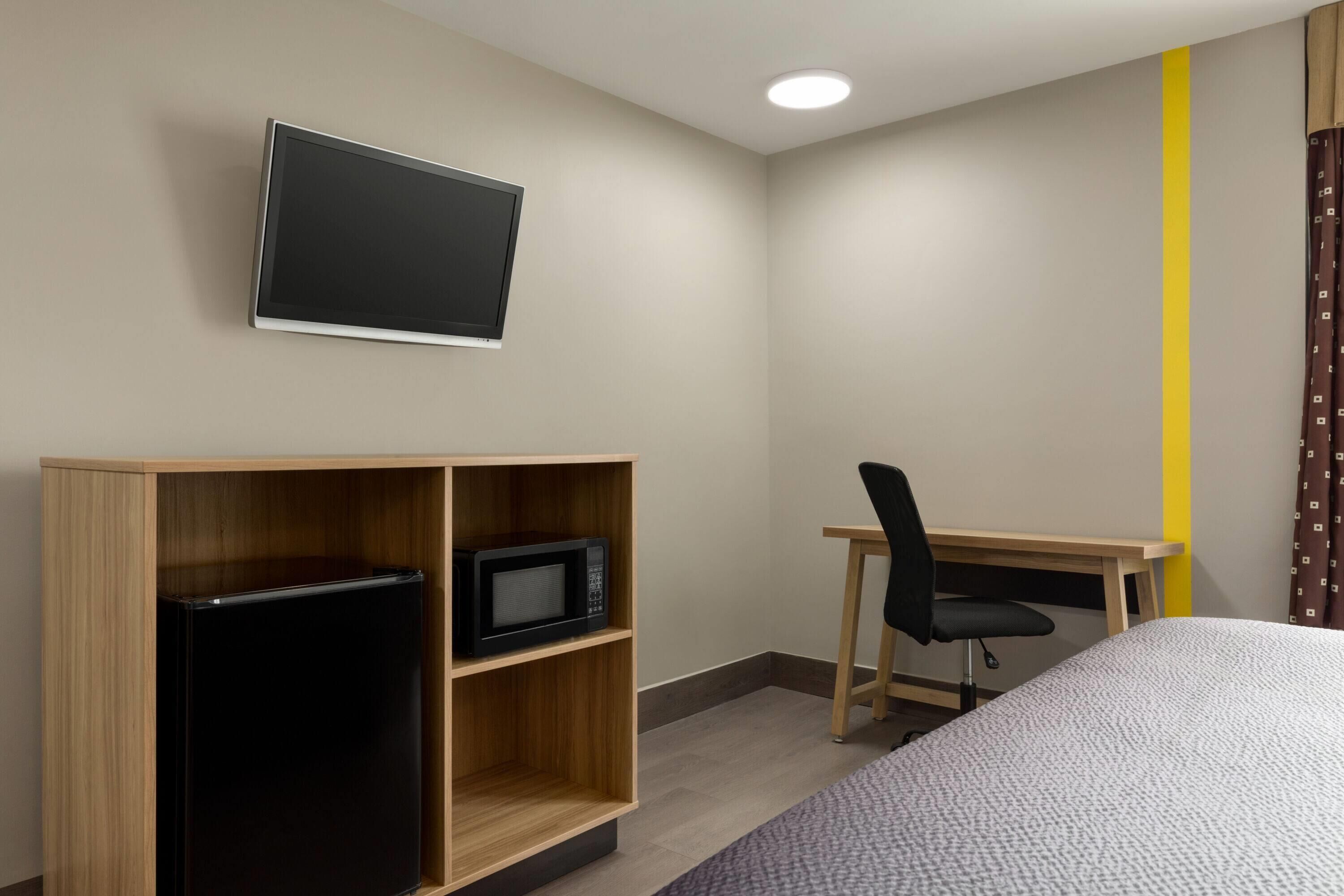 A hotel room with a bed, a desk with a chair, a TV mounted on the wall, and a refrigerator in a wooden cabinet.