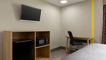 A hotel room with a bed, a desk with a chair, a TV mounted on the wall, and a refrigerator in a wooden cabinet.