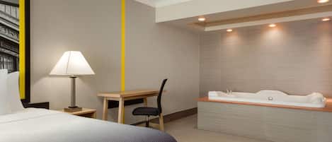 A hotel room with a bed, desk, chair, lamp, and a large bathtub.