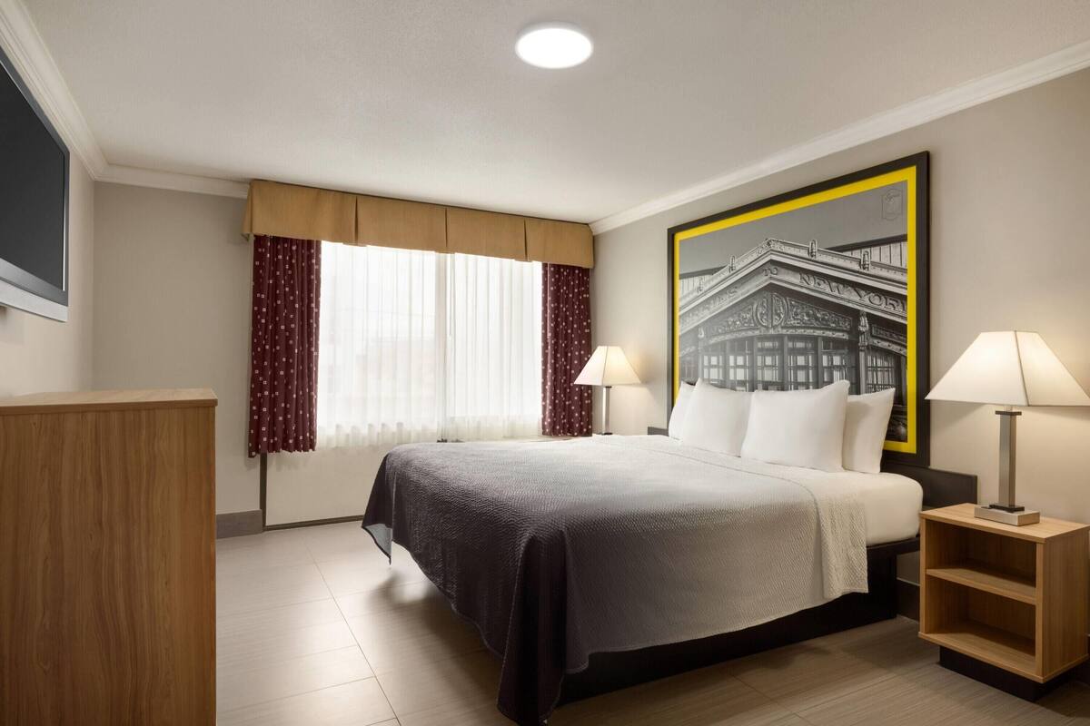 A hotel room with a large bed, two bedside tables with lamps, a wooden nightstand, a window with curtains, and a framed picture on the wall.