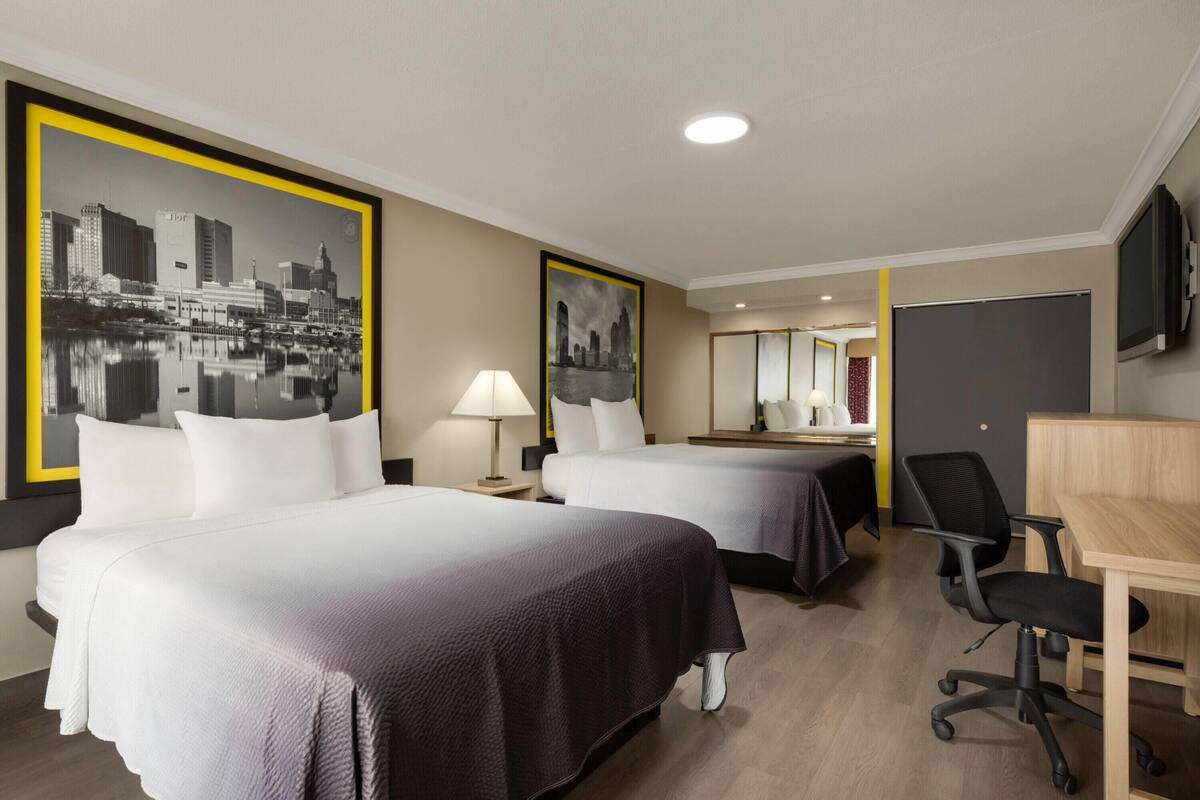 A hotel room with two beds, a desk, a chair, a TV, and a large cityscape painting on the wall.