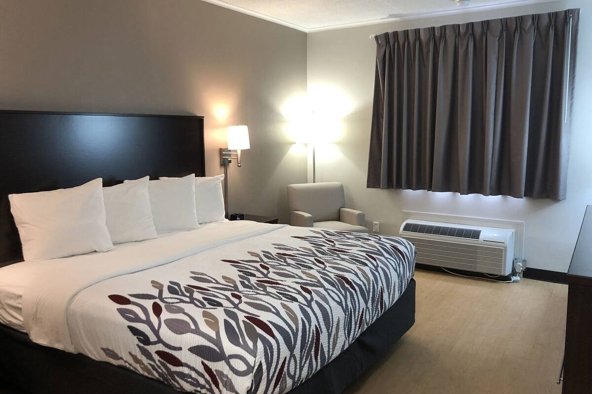 A hotel room with a large bed, a bedside table with a lamp, a chair, a window with curtains, and an air conditioning unit.