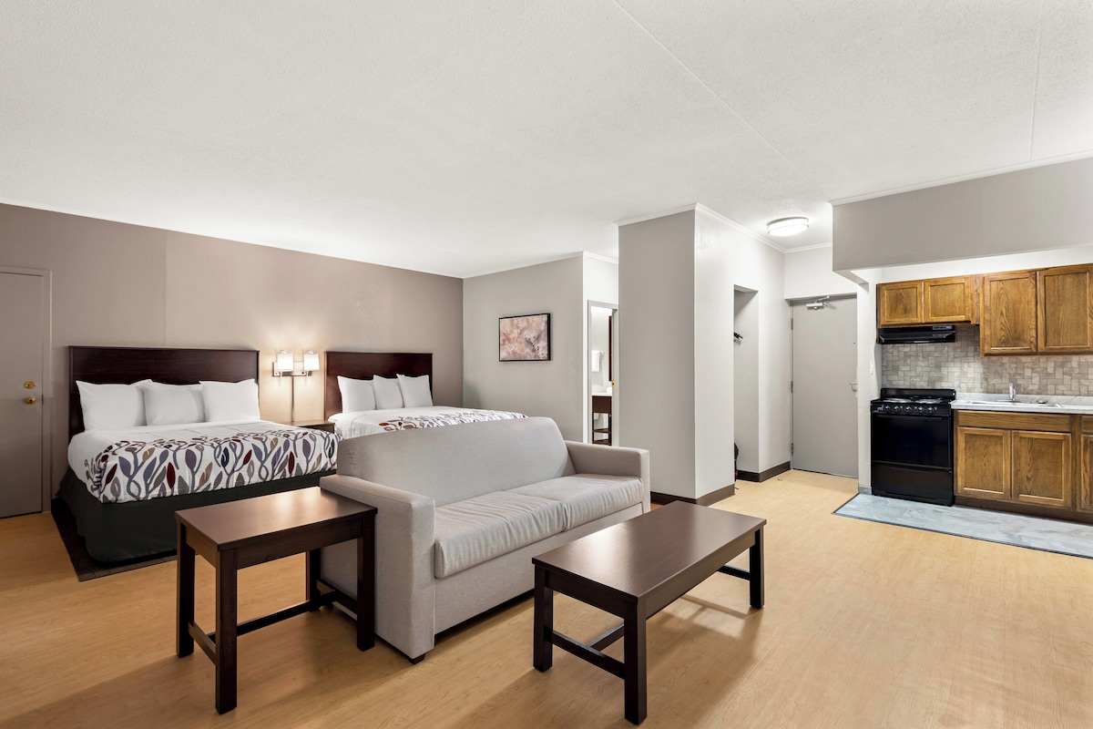 A modern hotel room with a bed, two sofas, a dining table, and a kitchenette.