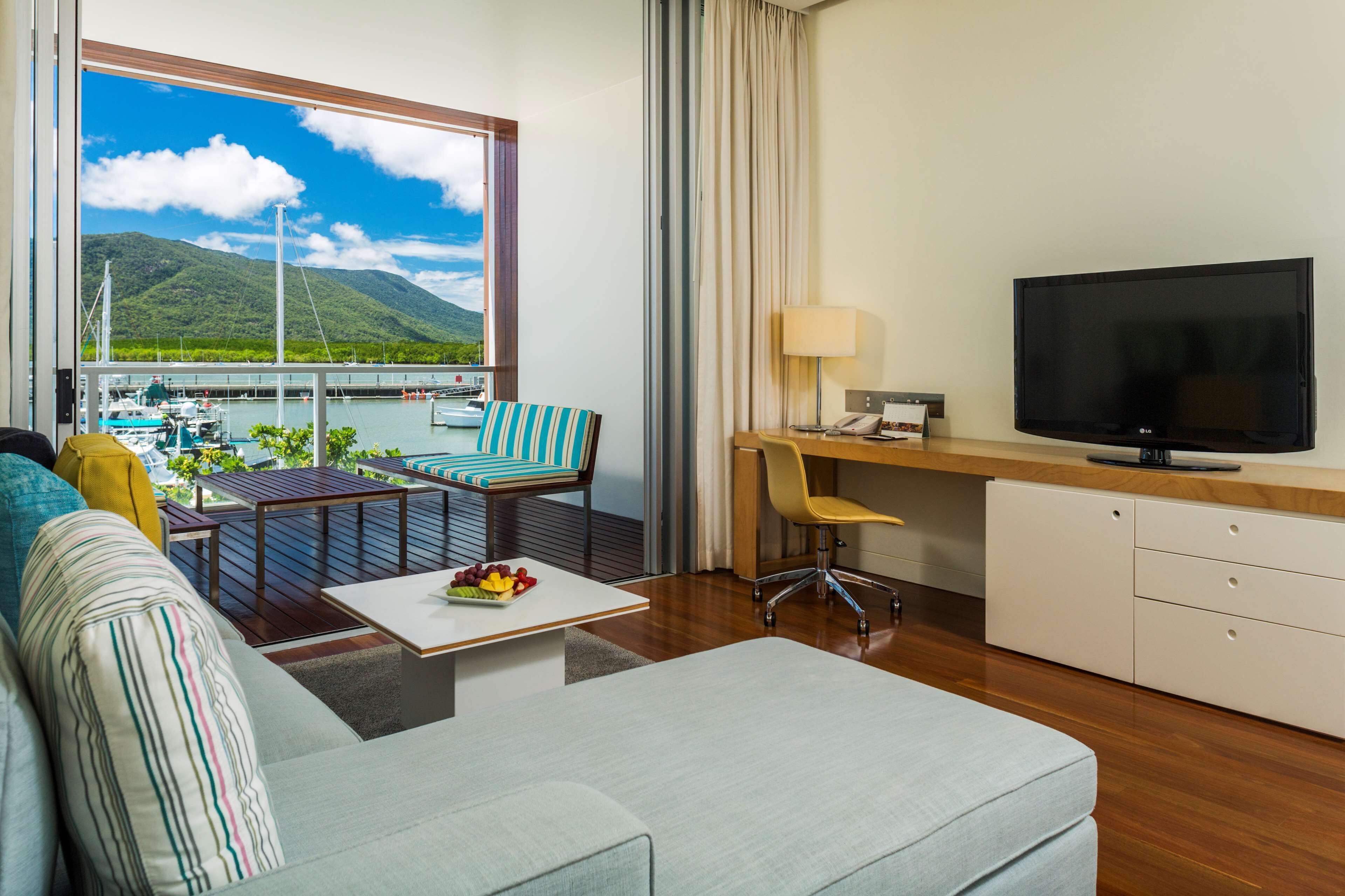 A modern hotel room with a large window offering a view of a marina and mountains. It includes a sofa, a dining table with fruit, a TV, and a desk with a chair.
