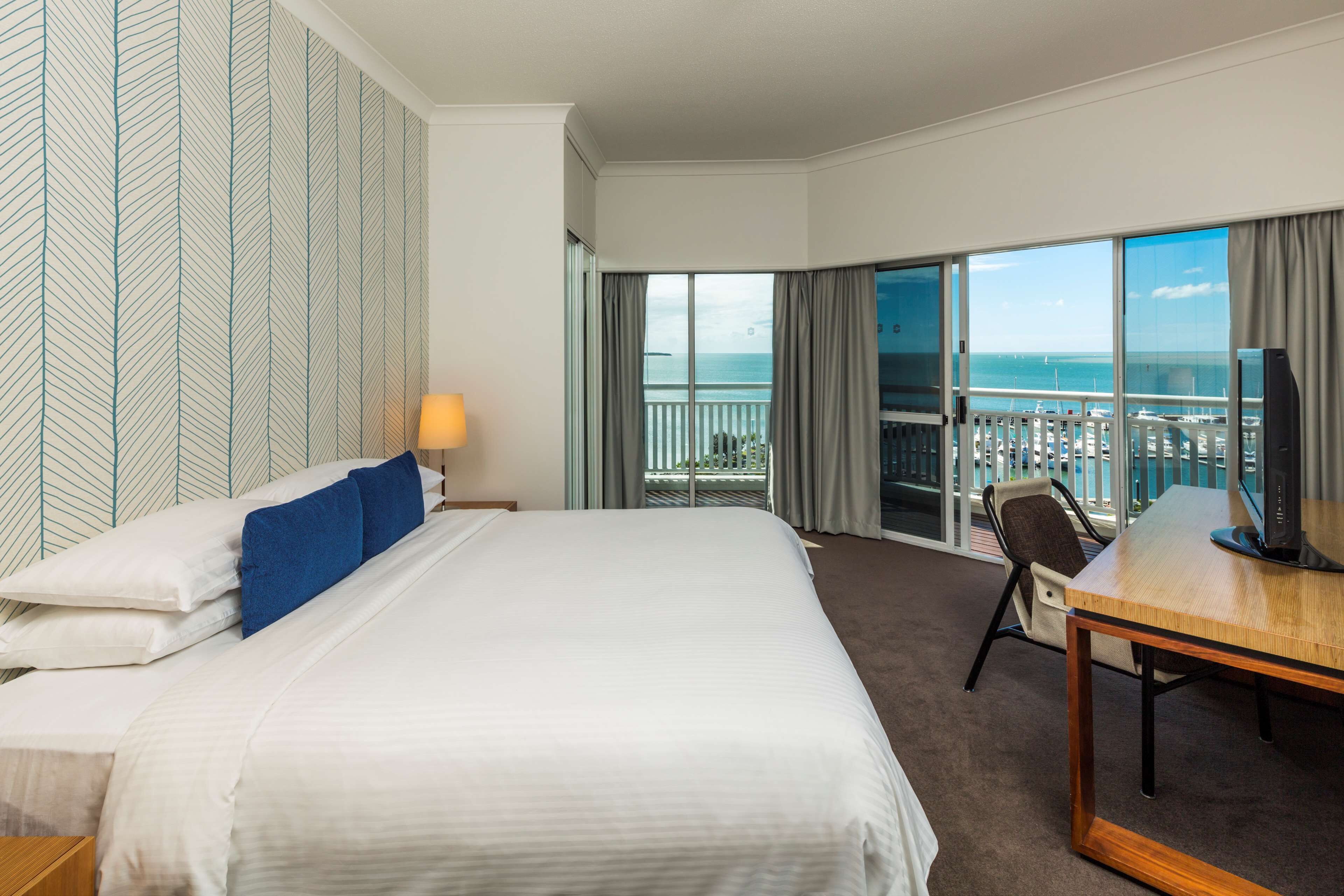 A hotel room with a large bed, a desk, a TV, and a balcony with a view of the sea.