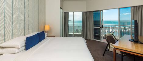 A hotel room with a large bed, a desk, a TV, and a balcony with a view of the sea.