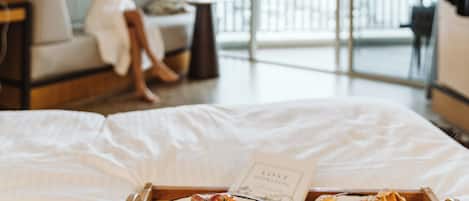 A hotel room with a bed, breakfast tray, and a person in a robe by the window.