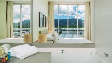 Une salle de bain d’hôtel moderne dotée d’une grande baignoire, d’un meuble-lavabo avec des articles de toilette, et offrant une vue sur une marina et des montagnes.