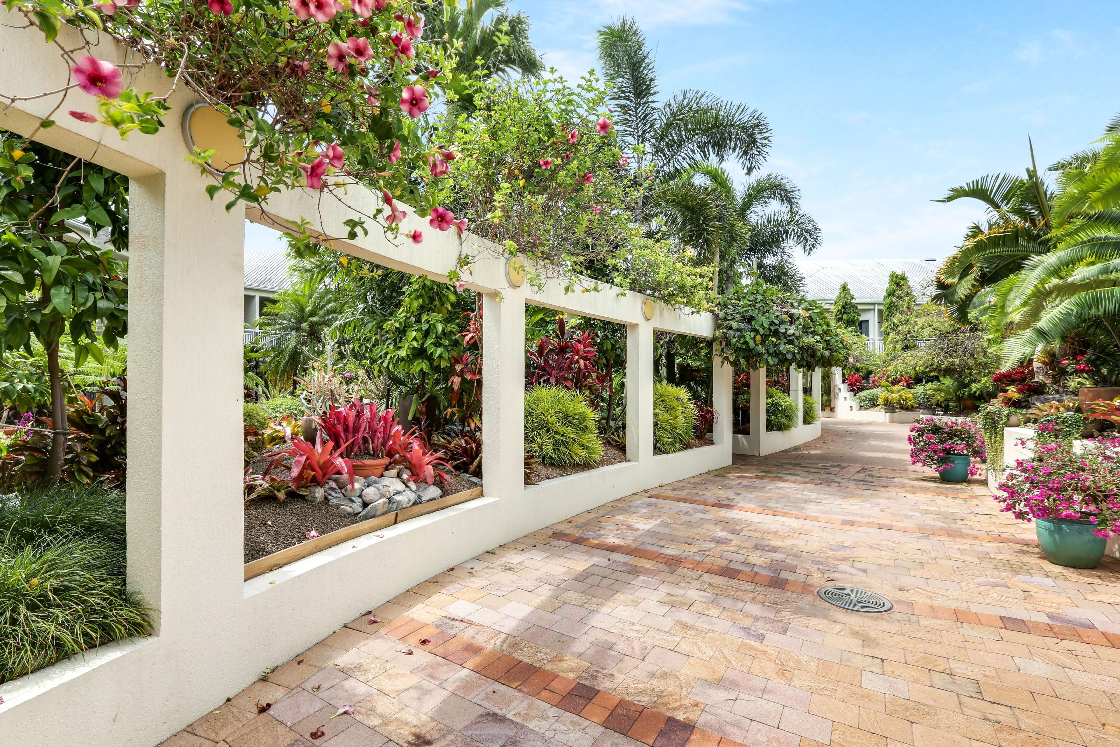A covered walkway with a brick floor, surrounded by lush greenery and flowering plants.