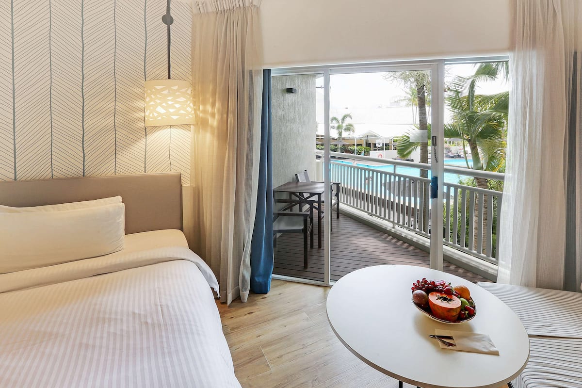 Habitación de hotel con cama, una mesa de comedor con fruta y un balcón con piscina.