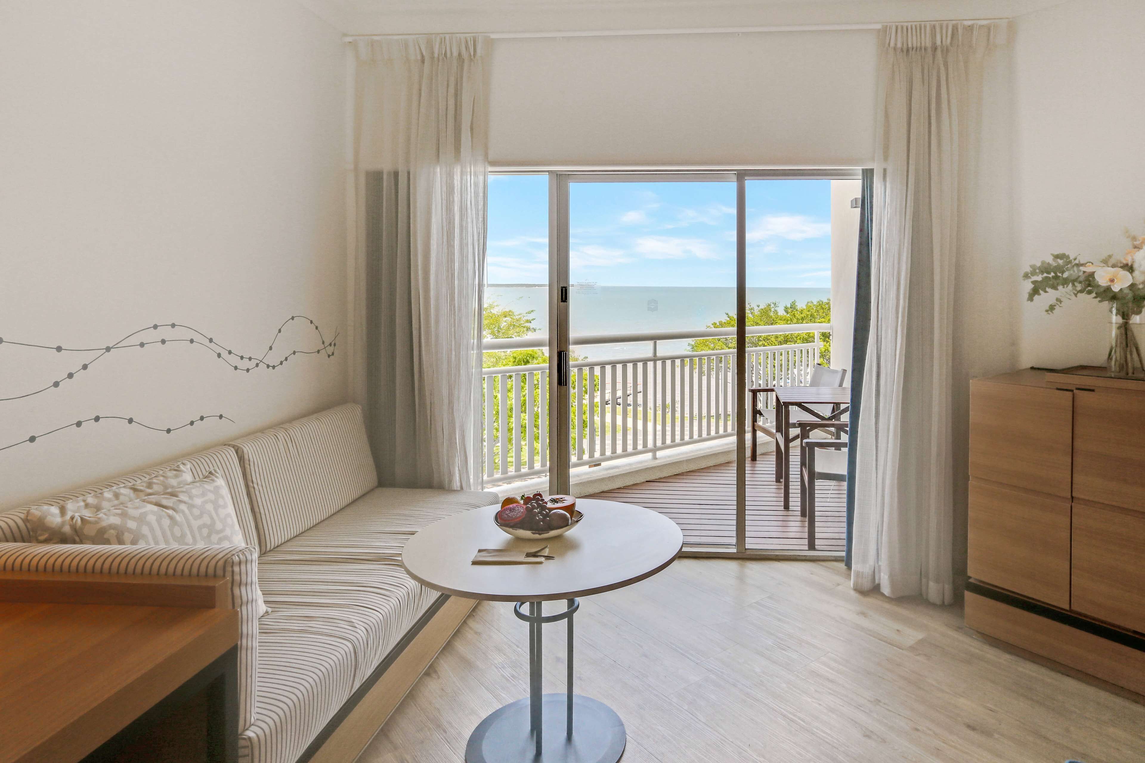 A room with a sofa, a round table with a bowl of fruit, a wooden cabinet, and a balcony with a view of the sea.