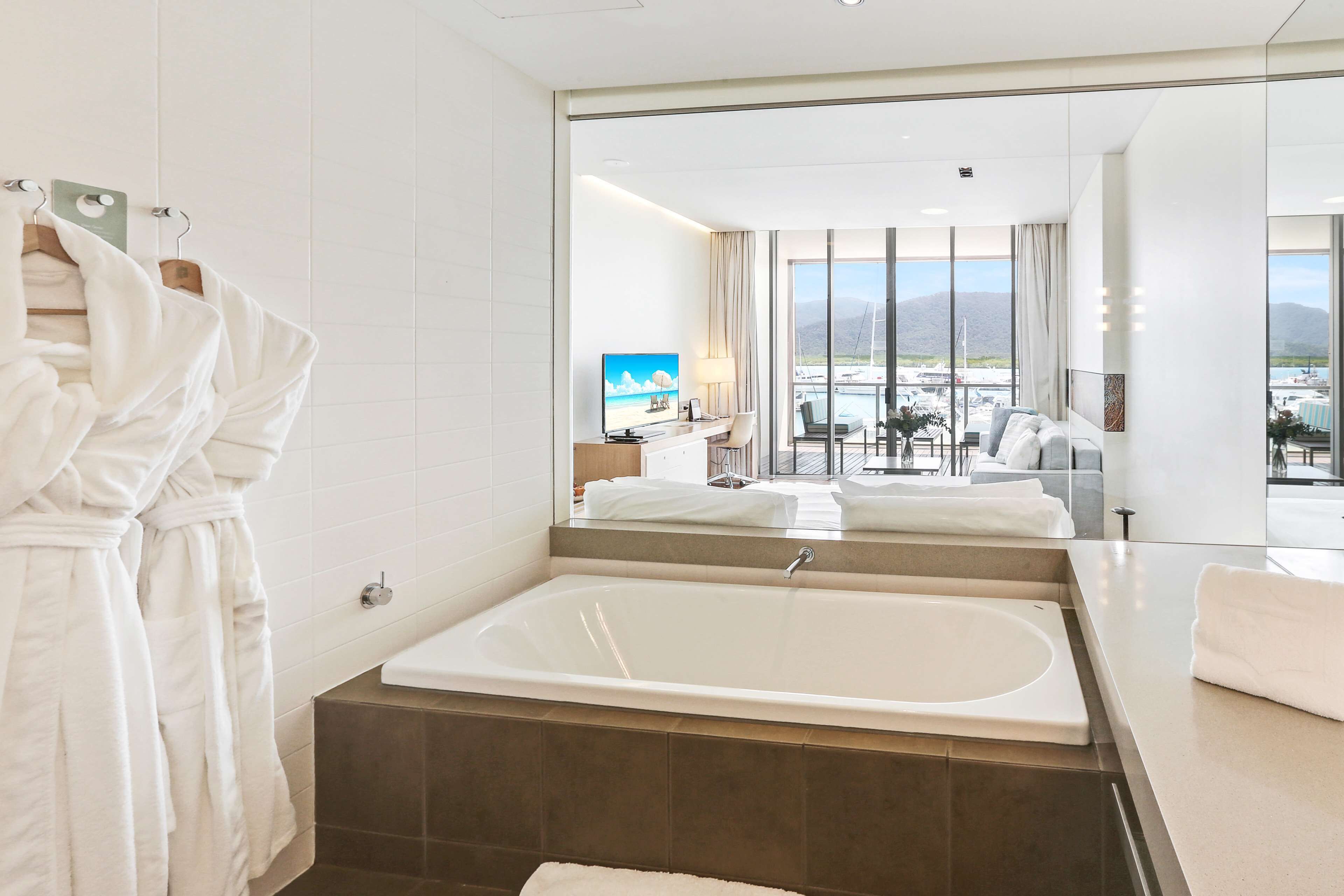 Ein modernes Badezimmer mit einer großen Badewanne, einem Spiegel, der einen Balkon mit Bergblick zeigt, und einem Satz weißer Bademäntel auf einem Wäscheständer.