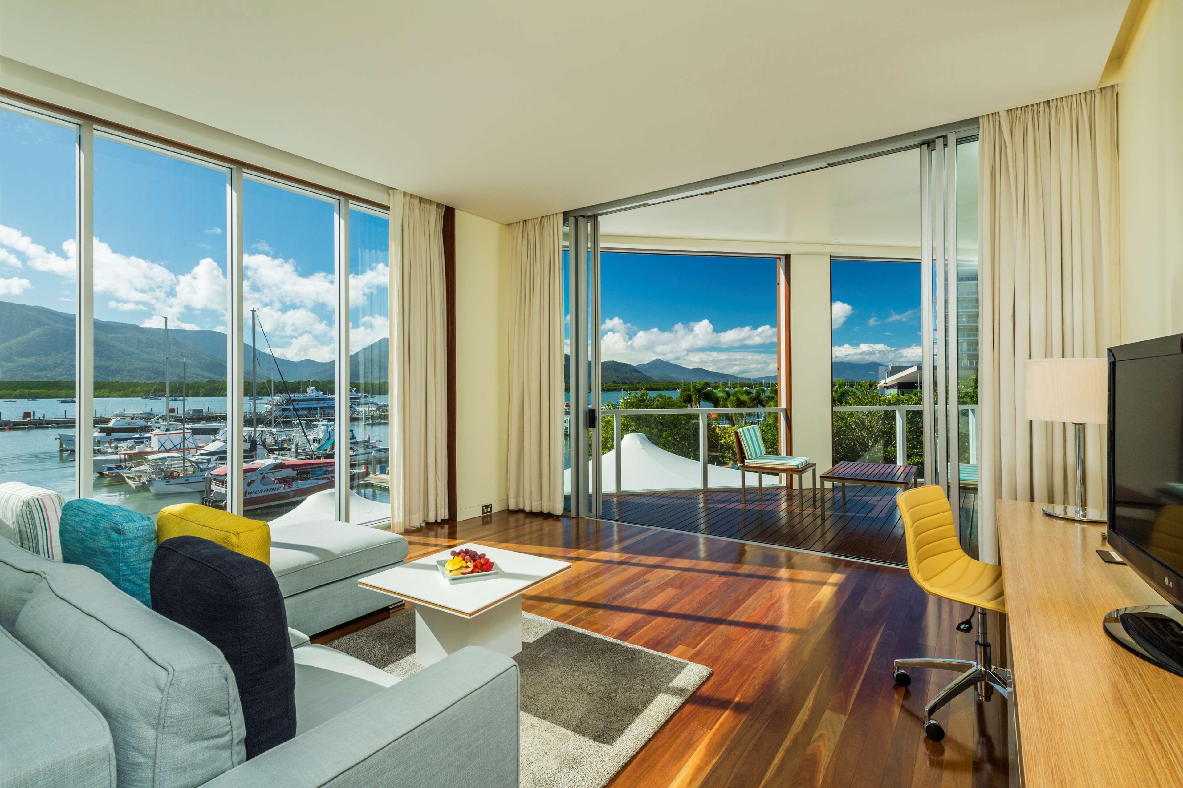 A modern hotel room with a large balcony, a sofa, a dining table, and a TV.
