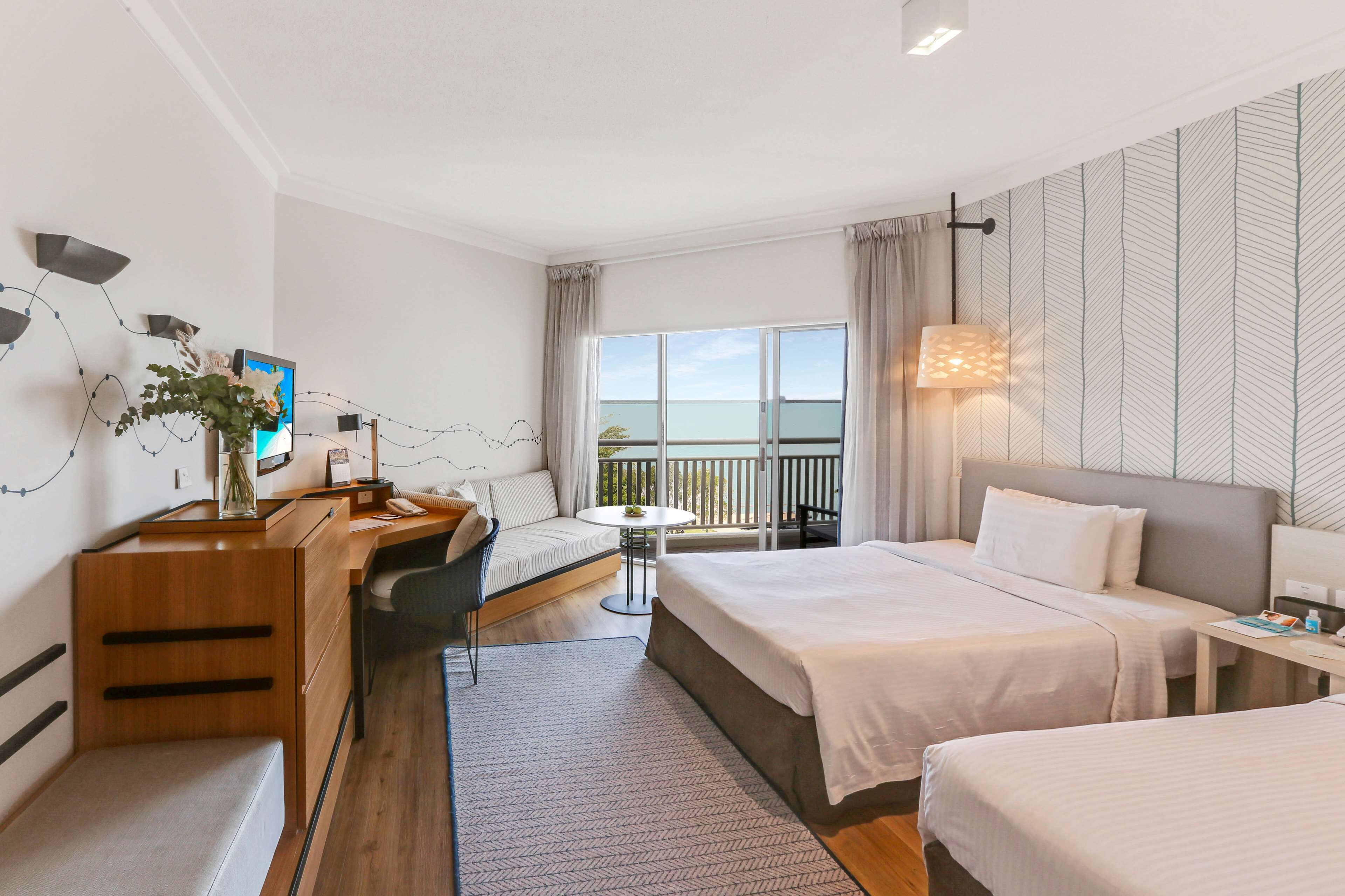 A hotel room with two beds, a desk, a chair, a small table, and a balcony with a view.