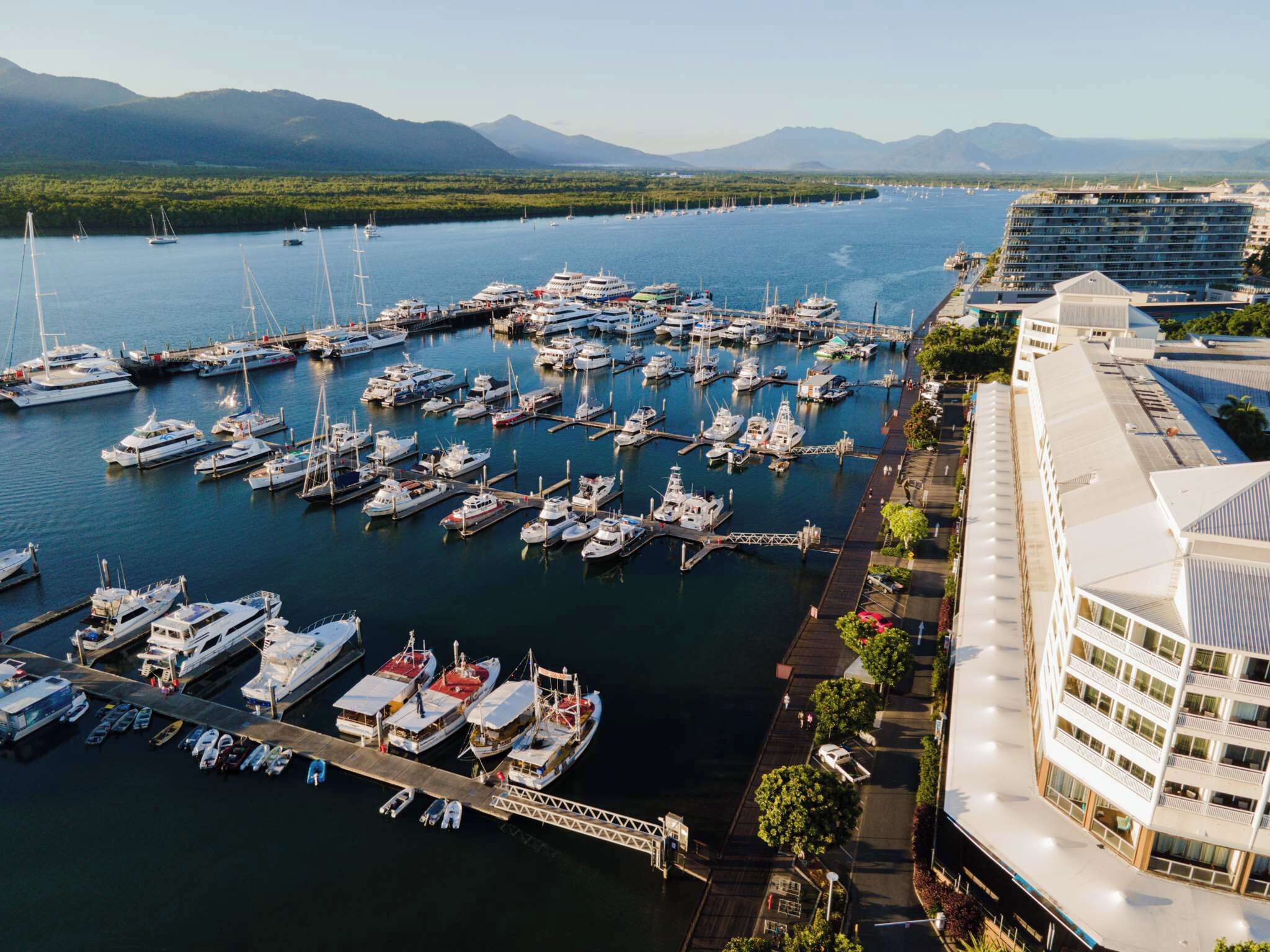 Un puerto deportivo con varios barcos amarrados, un edificio grande de varios pisos y un paisaje montañoso al fondo.
