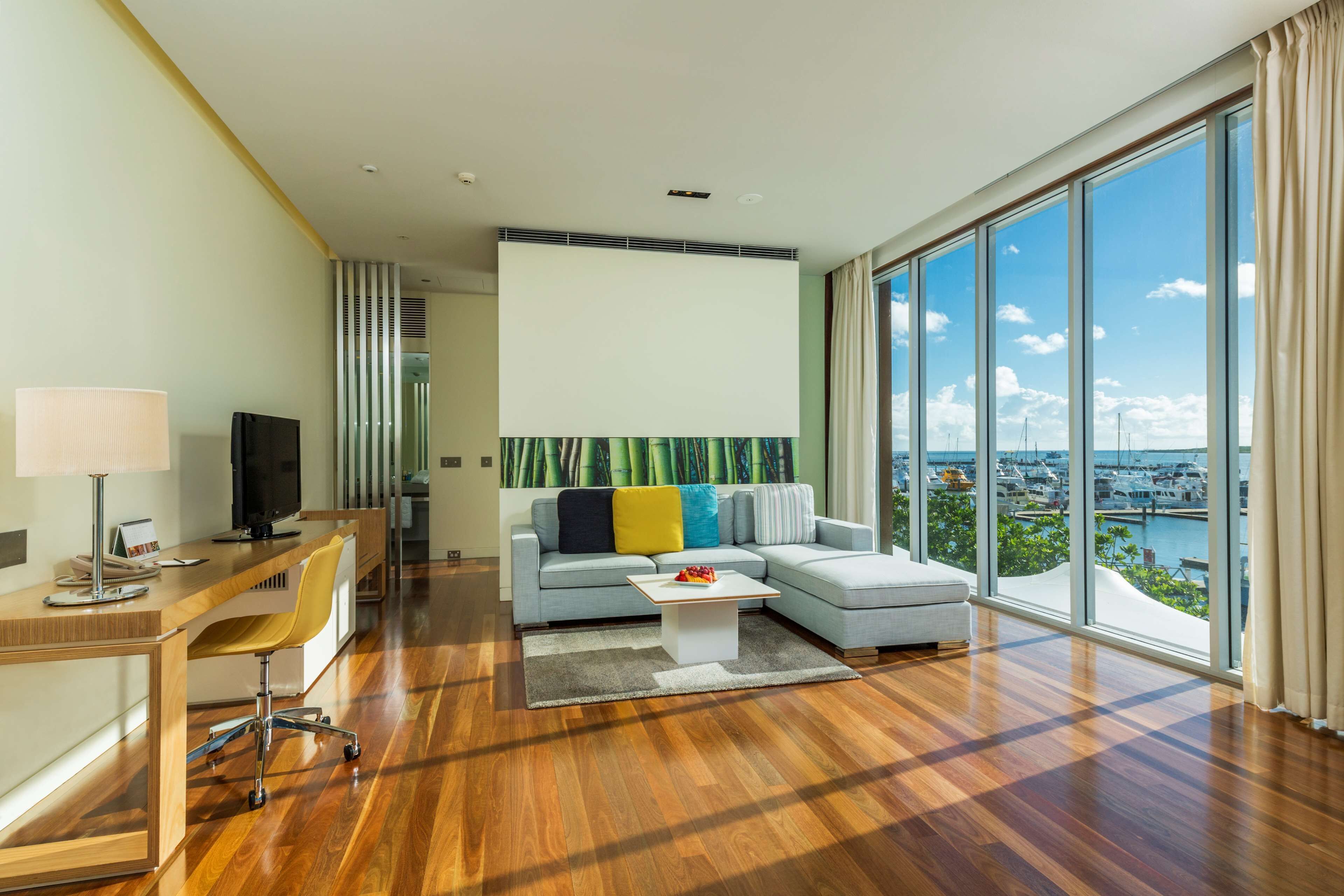 A modern living room with a large window, a sofa, a coffee table, and a desk with a lamp and a television.