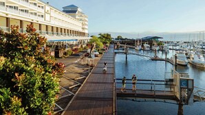 Un hotel frente al mar con una terraza de madera, un puerto deportivo con barcos y un edificio grande con balcones.