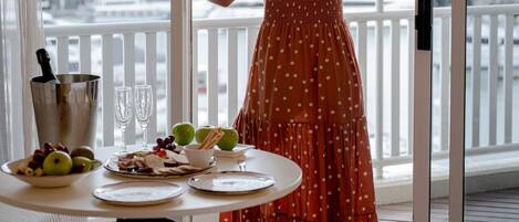 A woman in a polka dot dress and hat standing by a sliding glass door on a balcony overlooking a body of water and mountains.