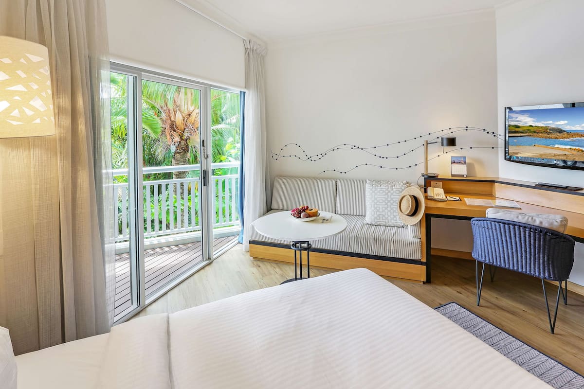 Habitación de hotel con cama, escritorio, silla, sofá, televisor y balcón con vistas a zonas verdes.