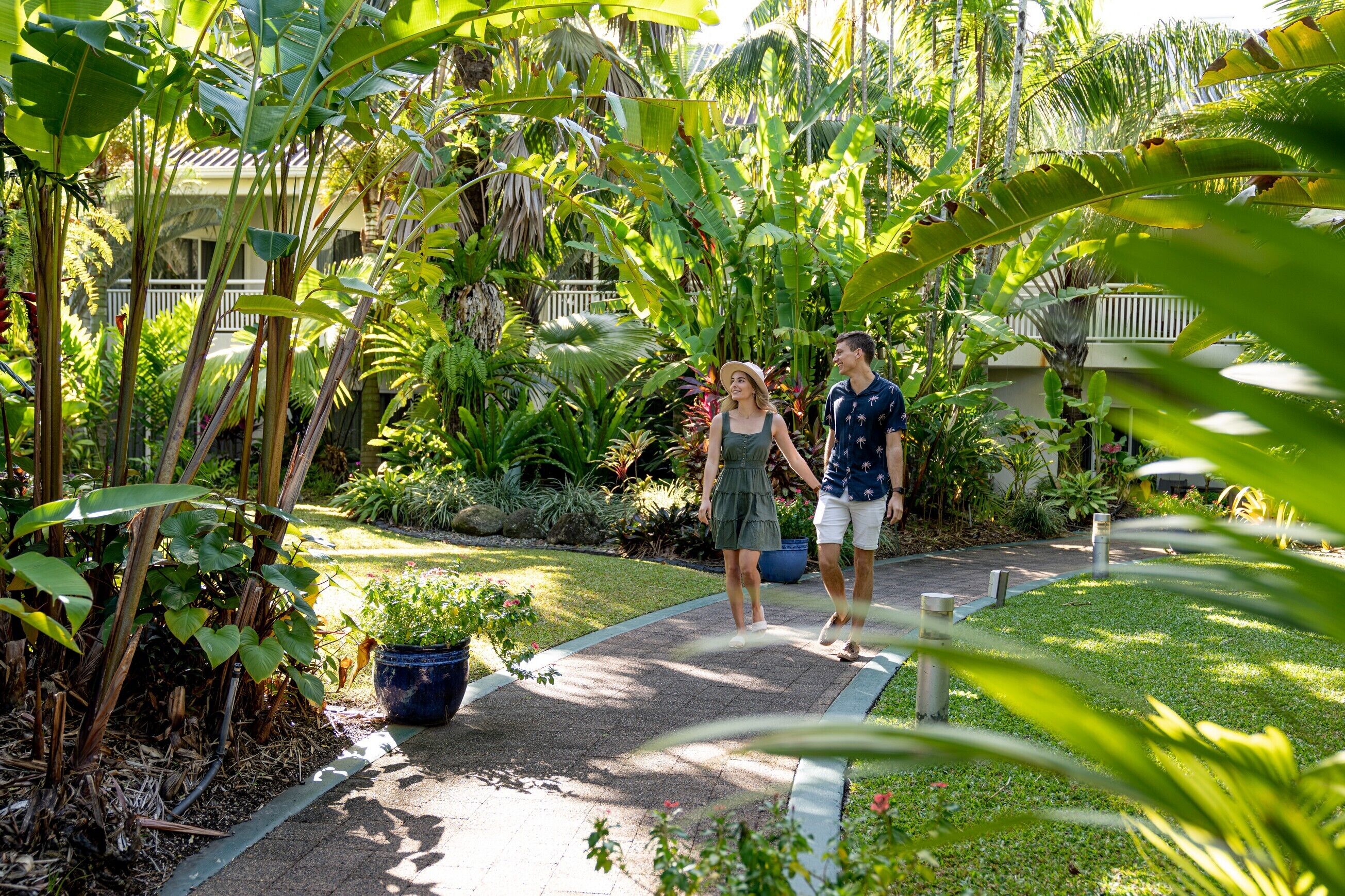 A pathway in a lush garden with various tropical plants and a white fence in the background.
