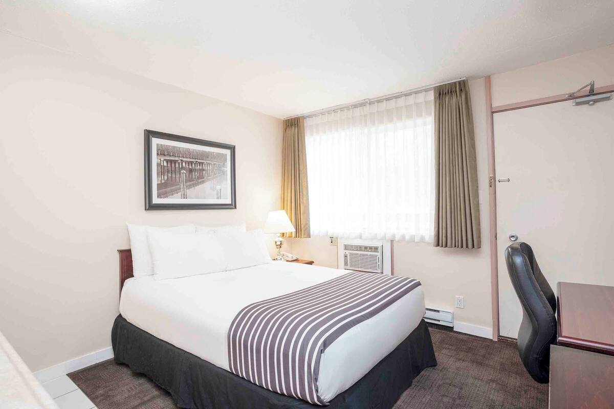 A hotel room with a bed, a desk, a chair, a lamp, a picture on the wall, and a window with curtains.