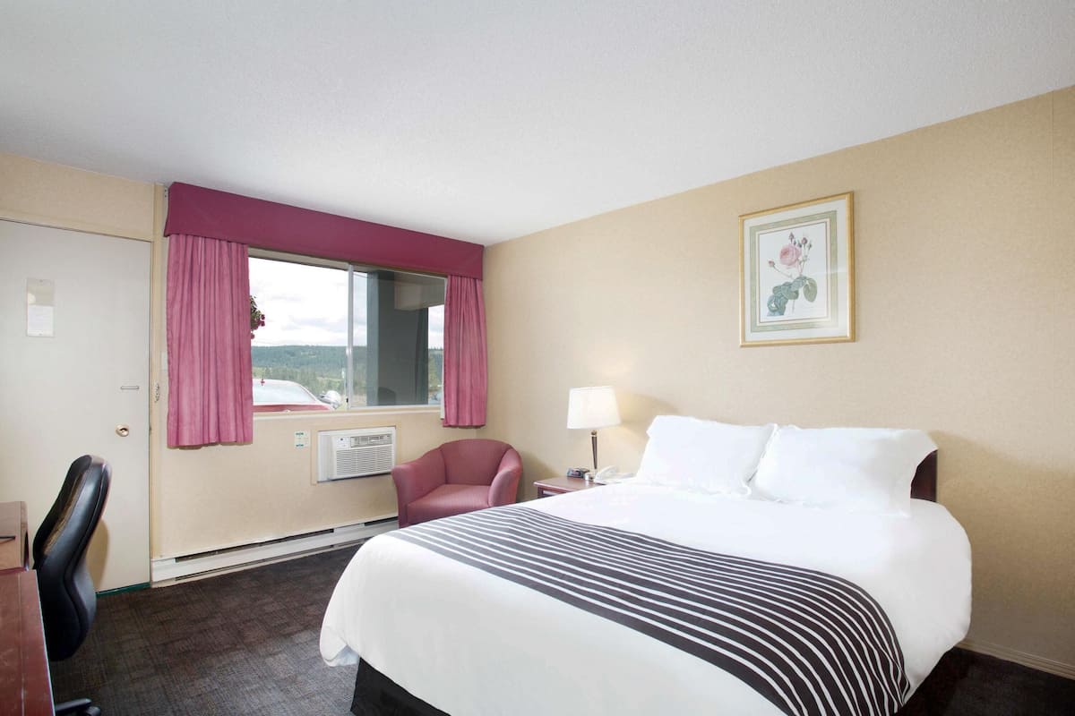 A hotel room with a bed, a desk, a chair, a window with curtains, a wall-mounted air conditioner, and a framed picture on the wall.