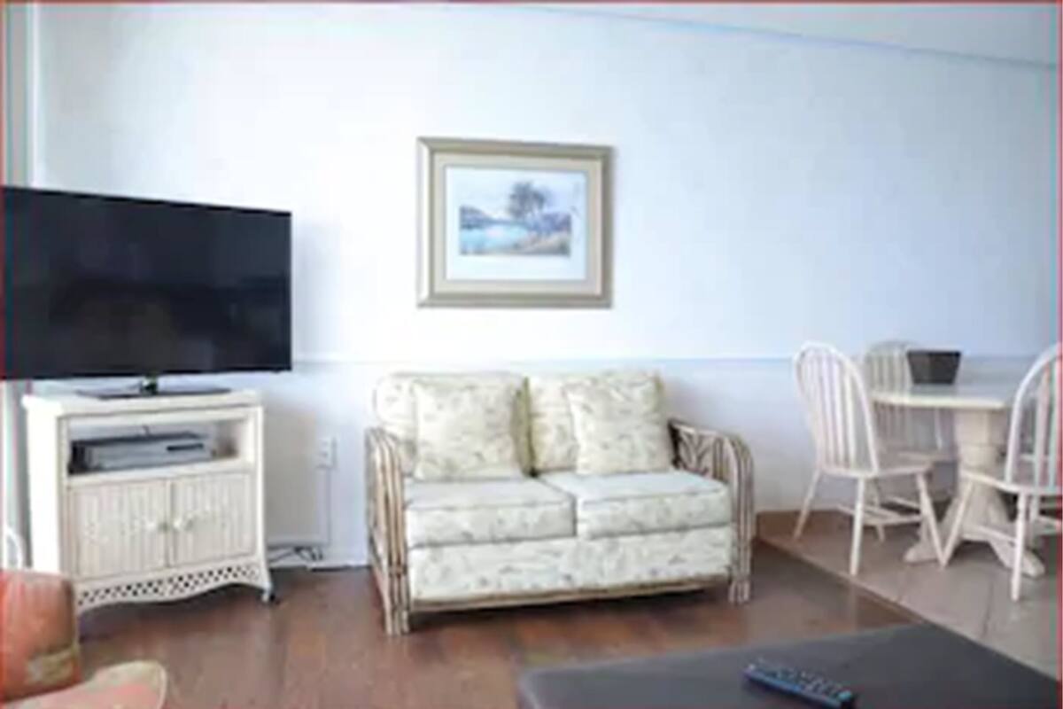 A living room with a sofa, a television on a stand, a framed picture on the wall, and a dining area with chairs.