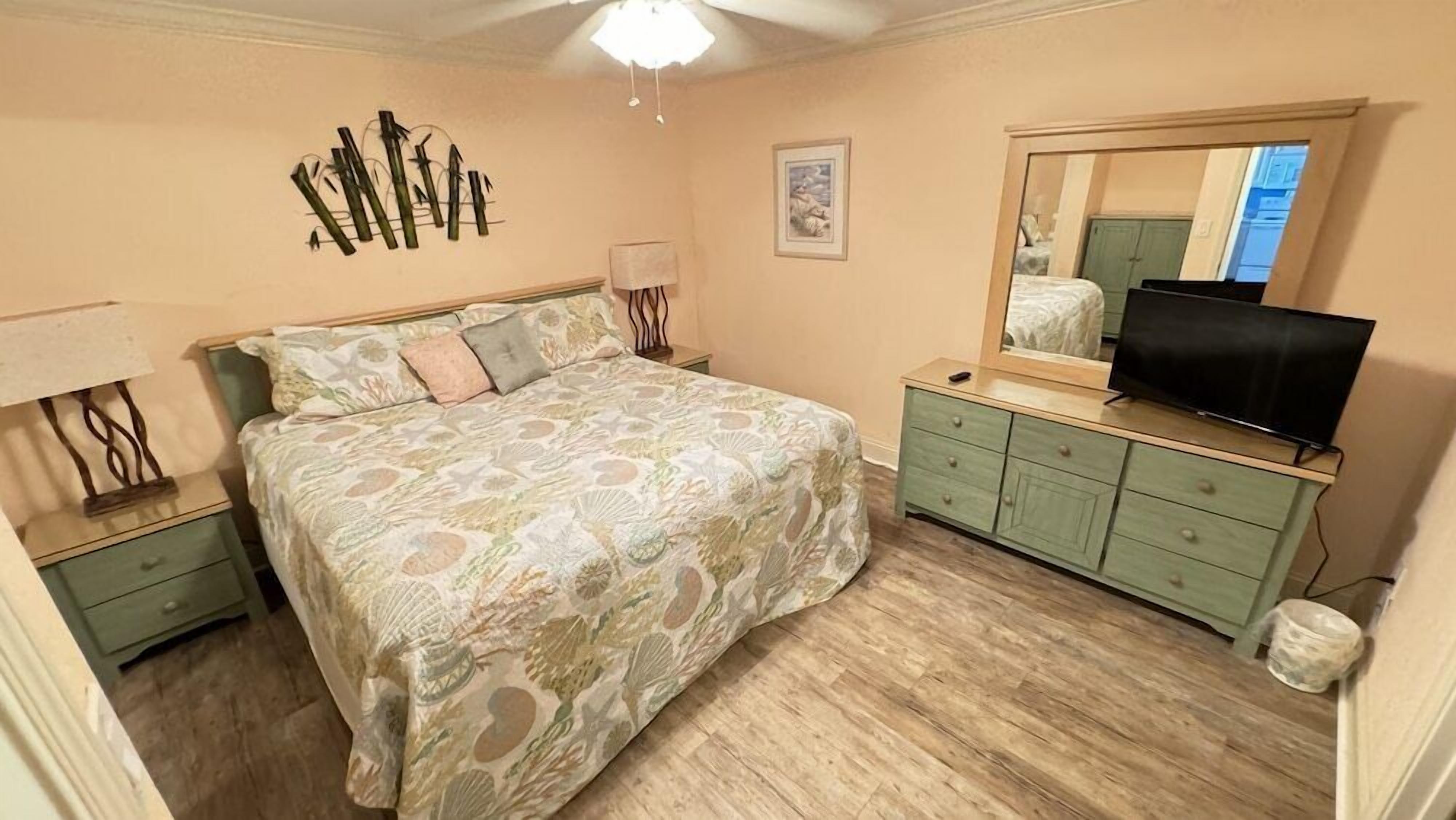 Quarto com cama, mesas de cabeceira, cômoda, televisão e espelho.