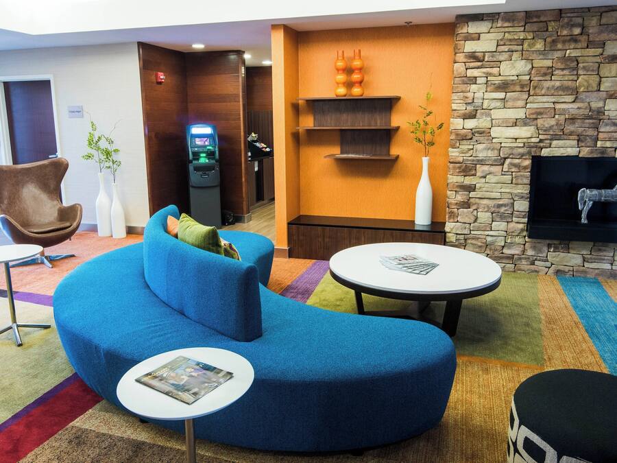 A modern hotel lobby with a blue sofa, a small round table, a TV mounted on a stone wall, and a reception desk with a digital kiosk.