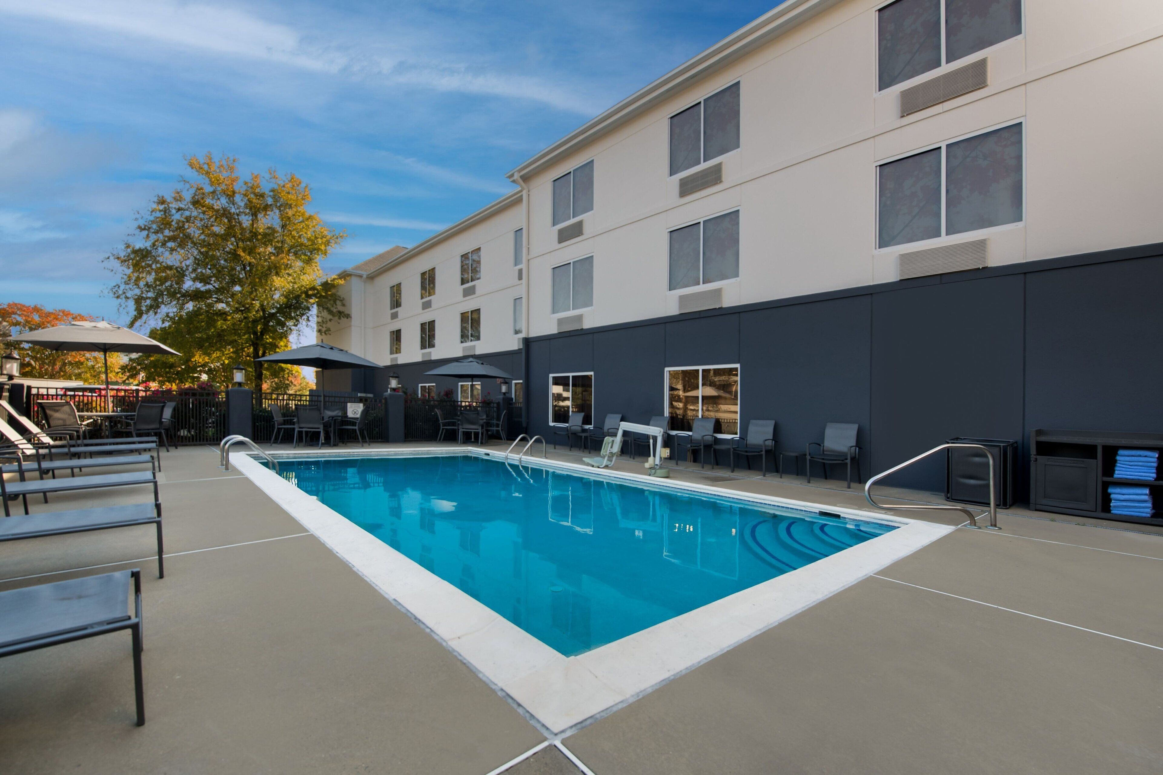A rectangular pool with a clear blue water surface, surrounded by lounge chairs and umbrellas, adjacent to a building with multiple windows and a dark facade.
