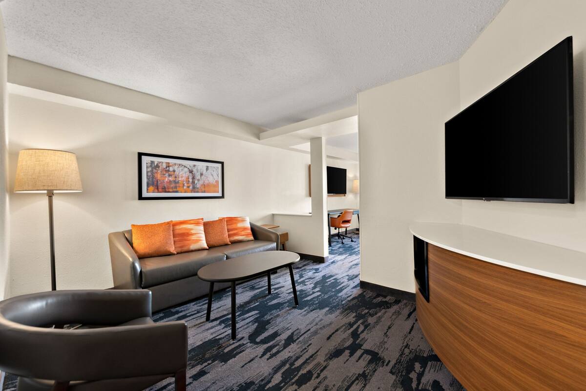 A modern hotel room with a sofa, armchair, coffee table, flat-screen TV, and a wooden sideboard.