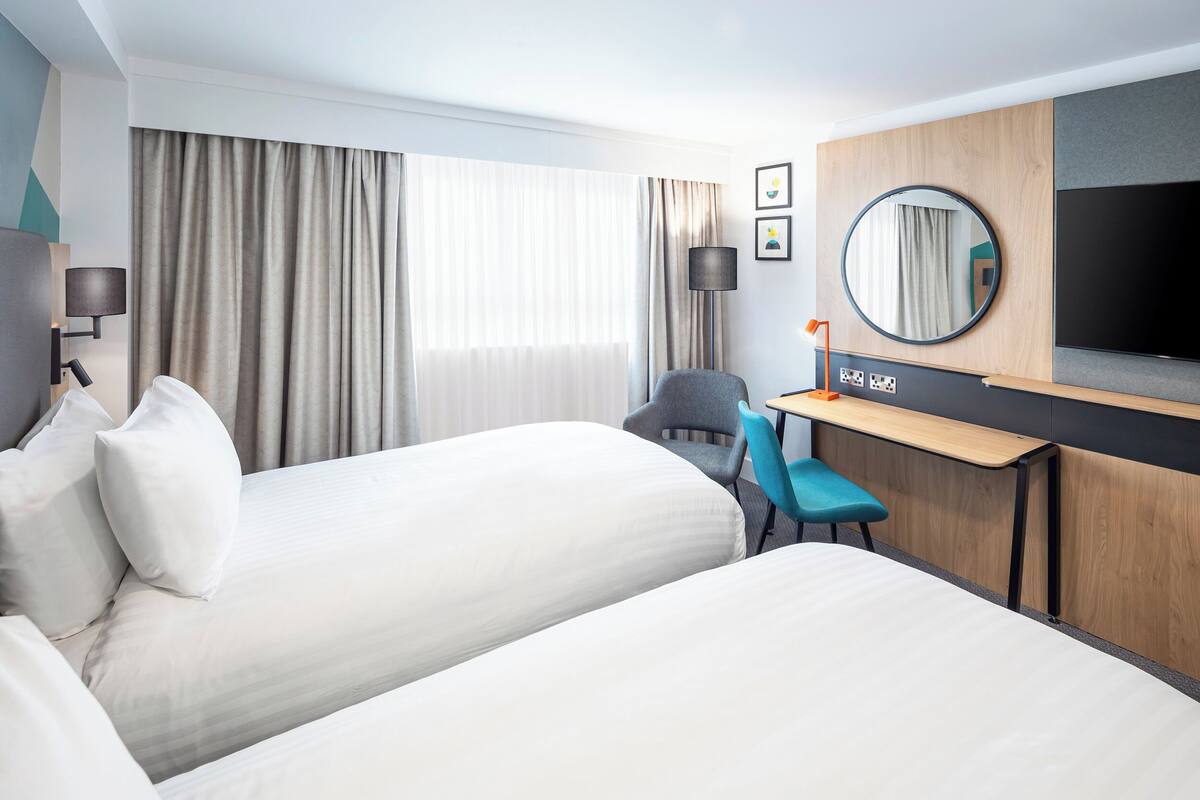 A hotel room with two beds, a desk with a computer, a chair, a TV, and a large window with curtains.