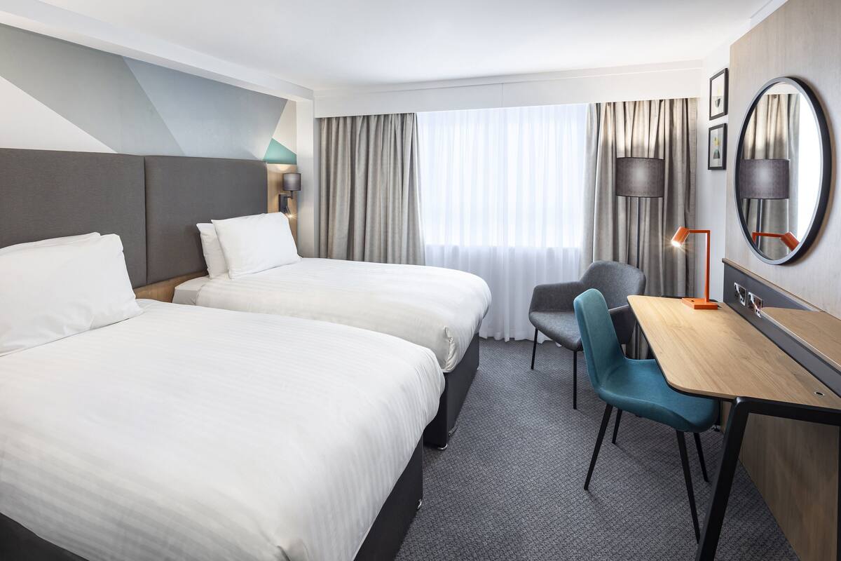 A hotel room with two beds, a desk, a chair, a lamp, and a large mirror.