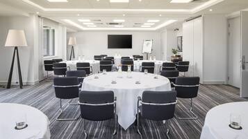 A conference room with a large round table, black chairs, and a white tablecloth. There are two standing lamps, a whiteboard, and a television on the wall.