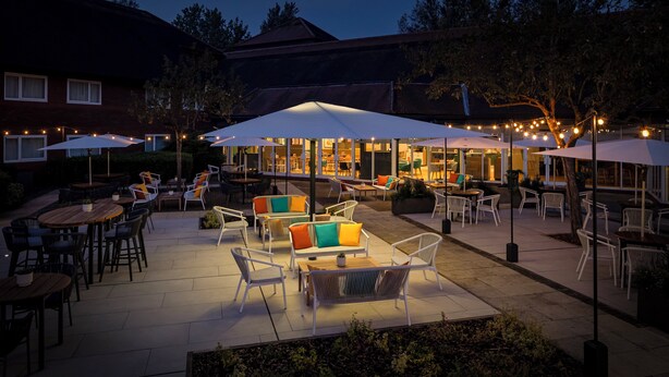 An outdoor patio area with tables and chairs, umbrellas, and a building in the background. - Holiday Inn Aylesbury by IHG (Aylesbury)