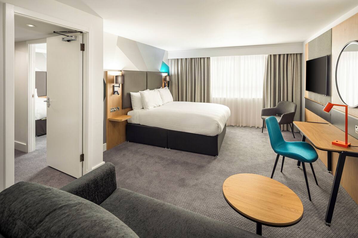 A modern hotel room with a large bed, a desk with a chair, a sofa, and a TV.
