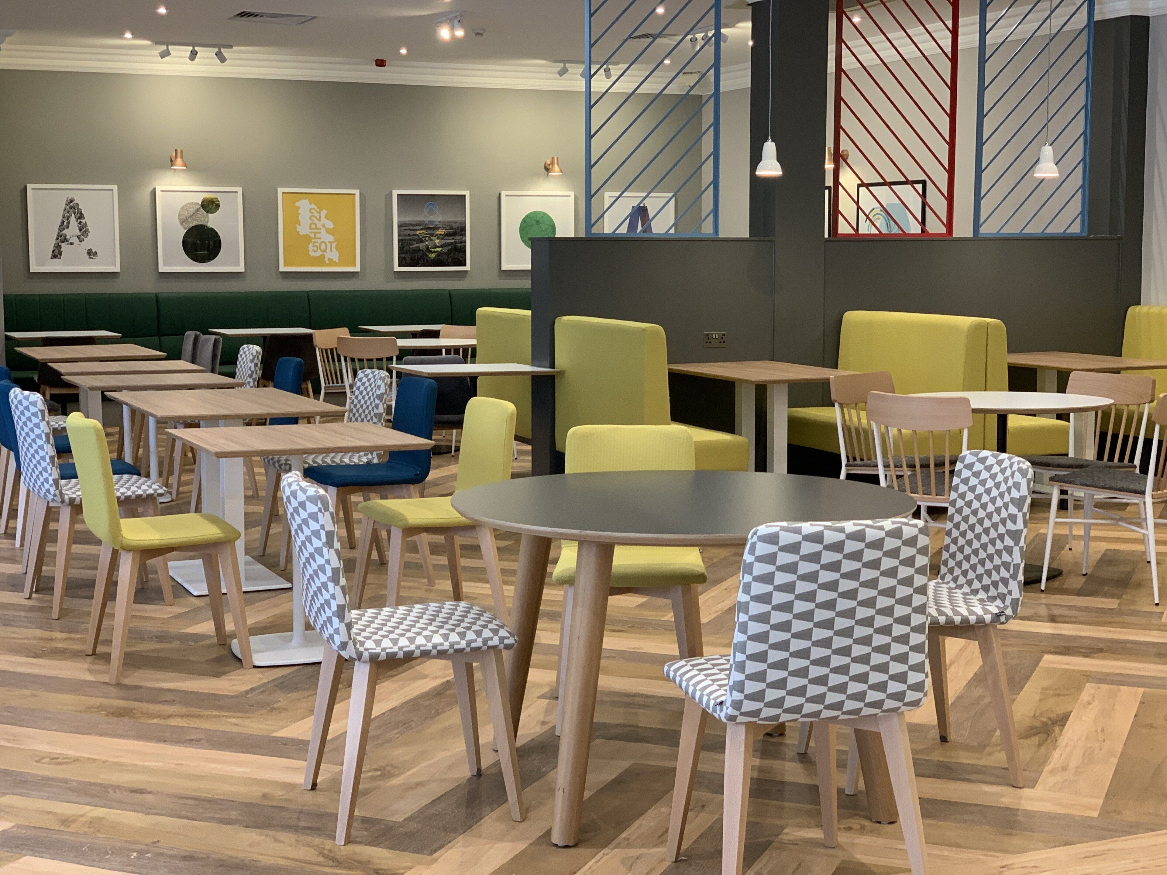 A modern cafe with wooden tables and chairs, a green cushioned seating area, and artwork on the walls.