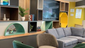 Ein modernes Wohnzimmer mit einem grauen Ecksofa, einem kleinen runden Tisch und einem grünen Sessel. An der Wand ist ein Flachbildfernseher angebracht, umgeben von Regalen mit Büchern und Dekoration.