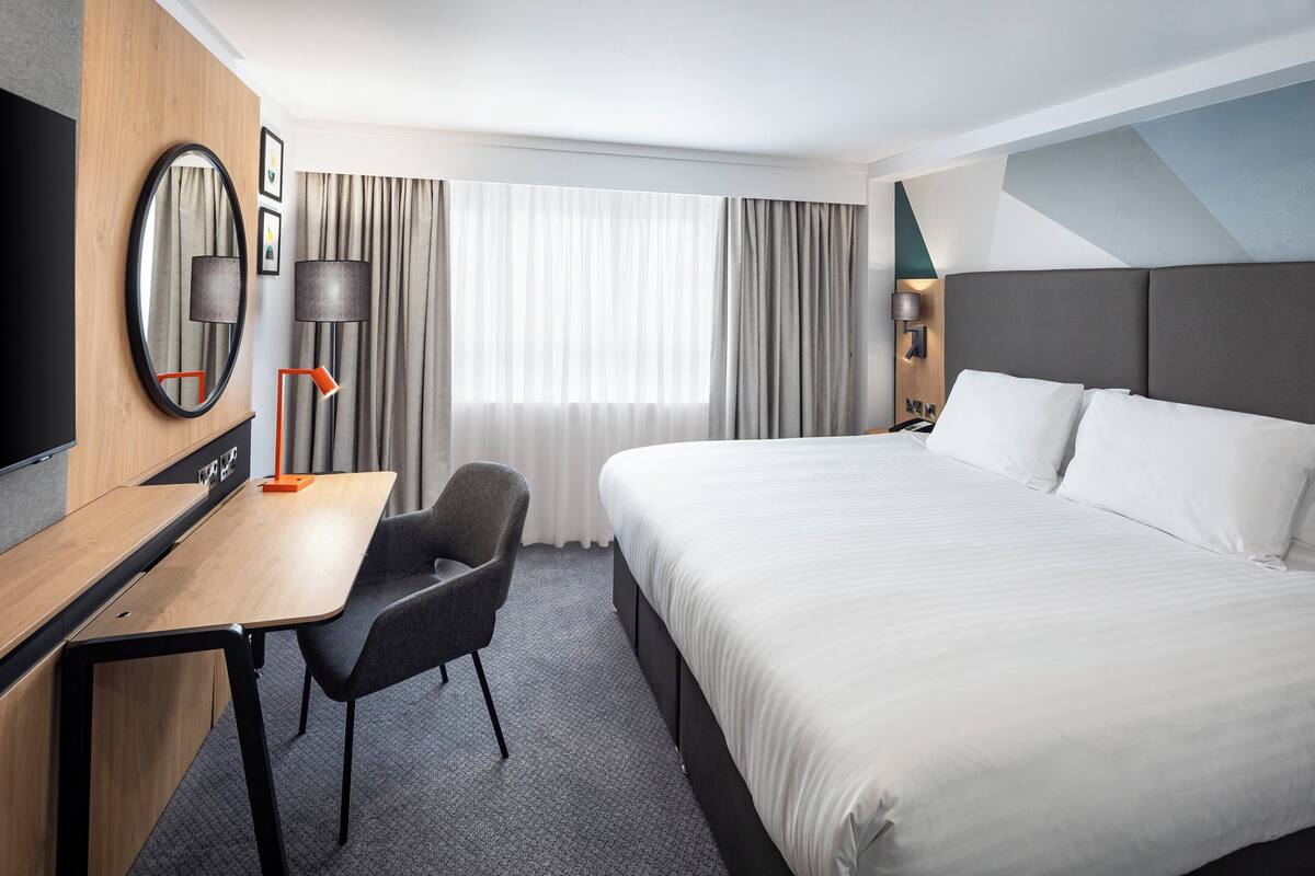 A hotel room with a bed, a desk, a chair, a TV, and a window with curtains.