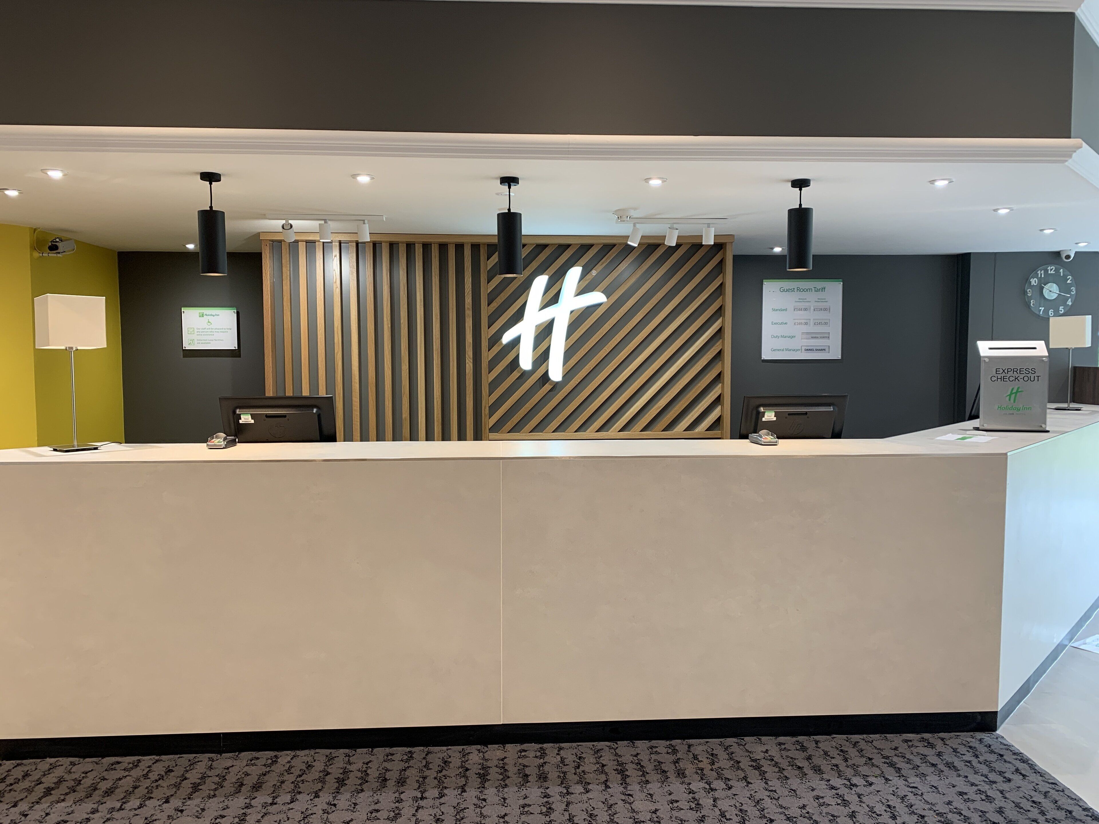 A hotel reception desk with a modern design, featuring a large illuminated 'H' logo, a digital guest room tariff display, and an express check-out sign.